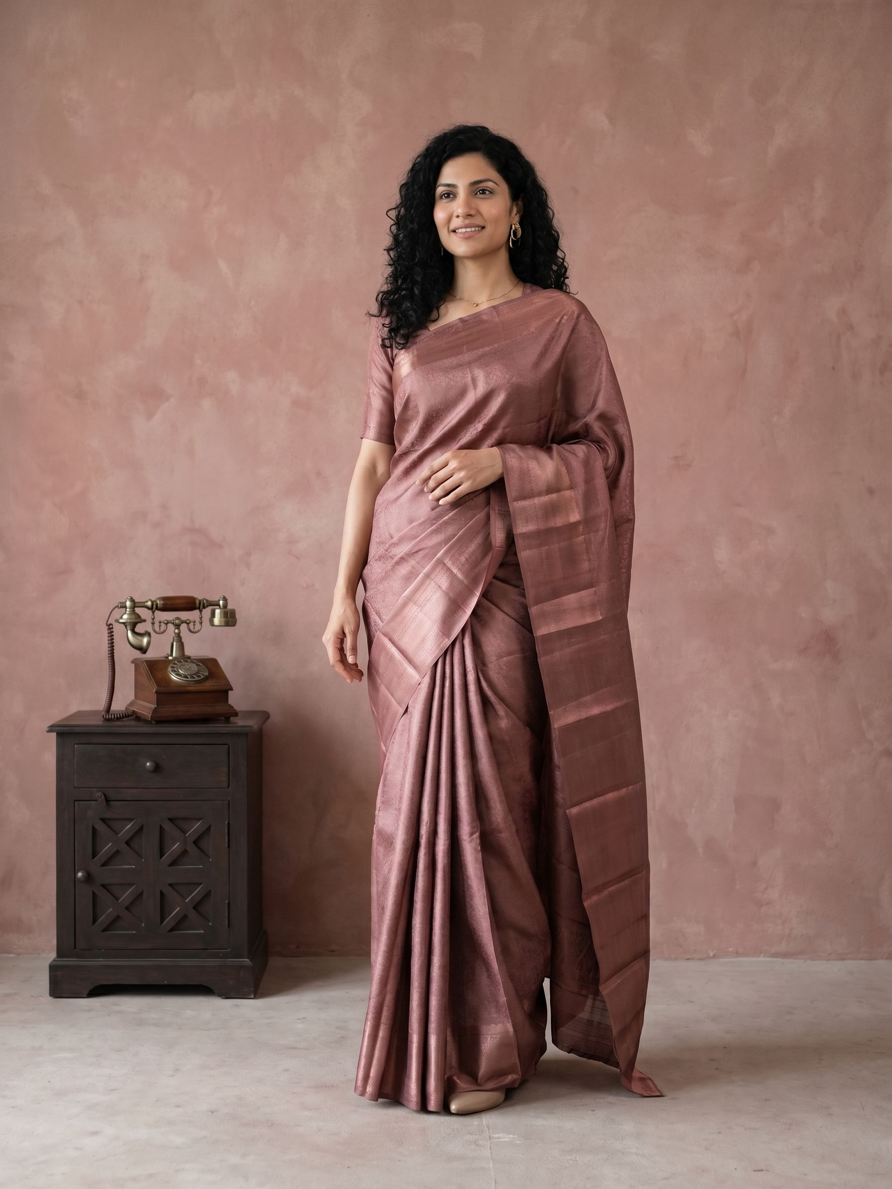 Woman in a brown saree standing next to a vintage telephone against a pink wall.