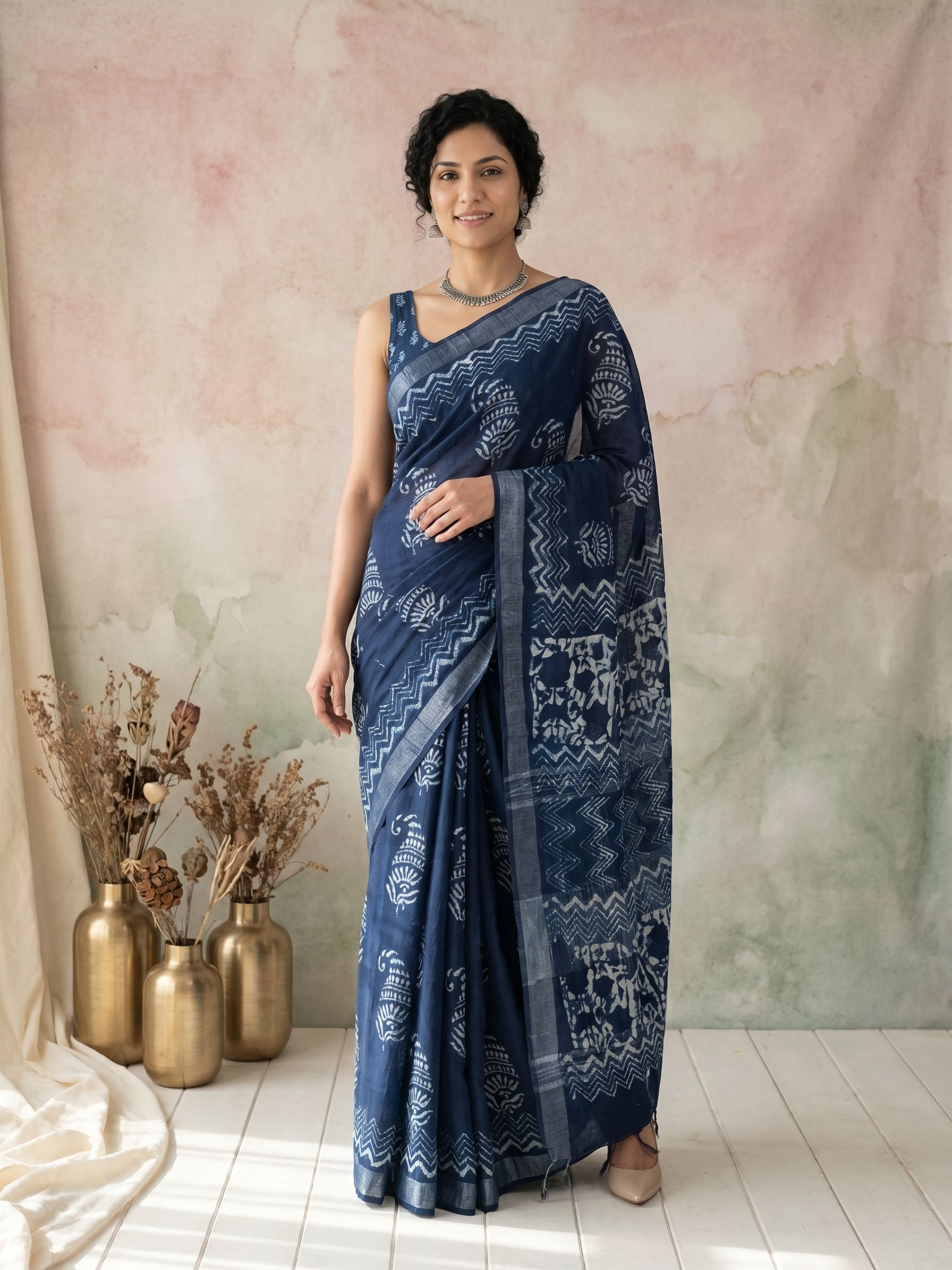 Woman wearing a blue saree with a pattern, standing against a soft pastel-colored wall.