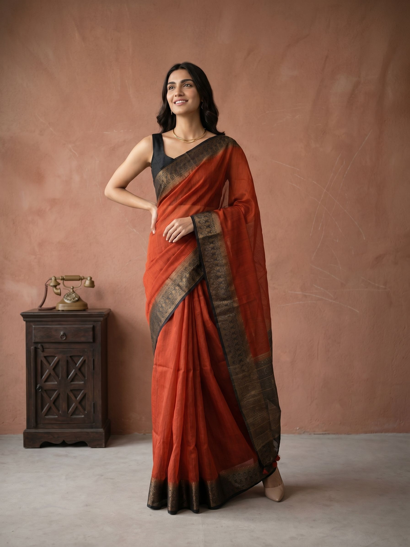 Woman in a red saree with black border standing against a brown wall.