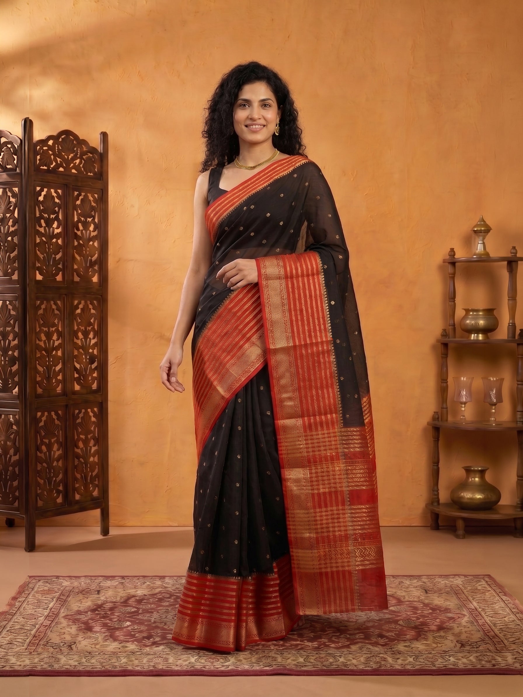 Woman wearing a black and red saree standing in a room with wooden furniture and a rug.