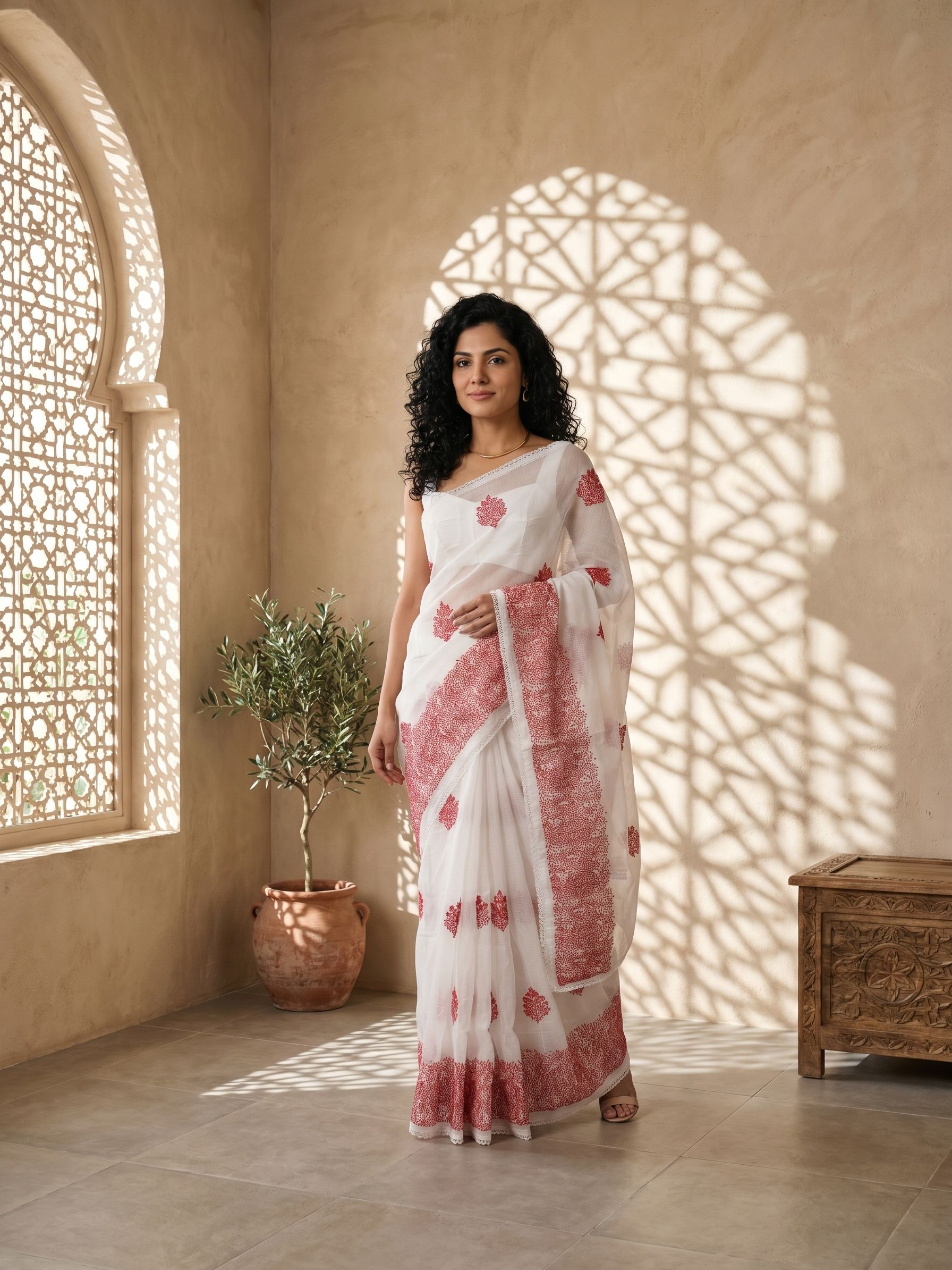 Woman in a white and red saree standing in a sunlit room with arched windows.
