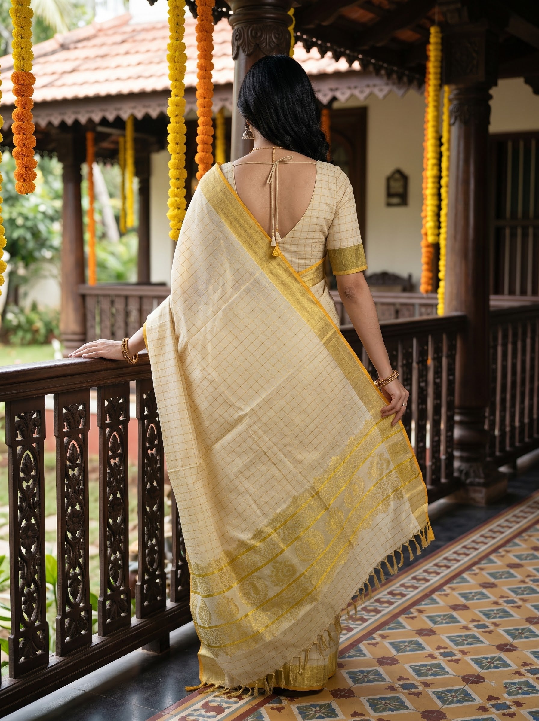 A woman wearing a light beige and yellow checkered saree with a yellow border and matching blouse, standing on a traditional wooden porch with marigold garlands.