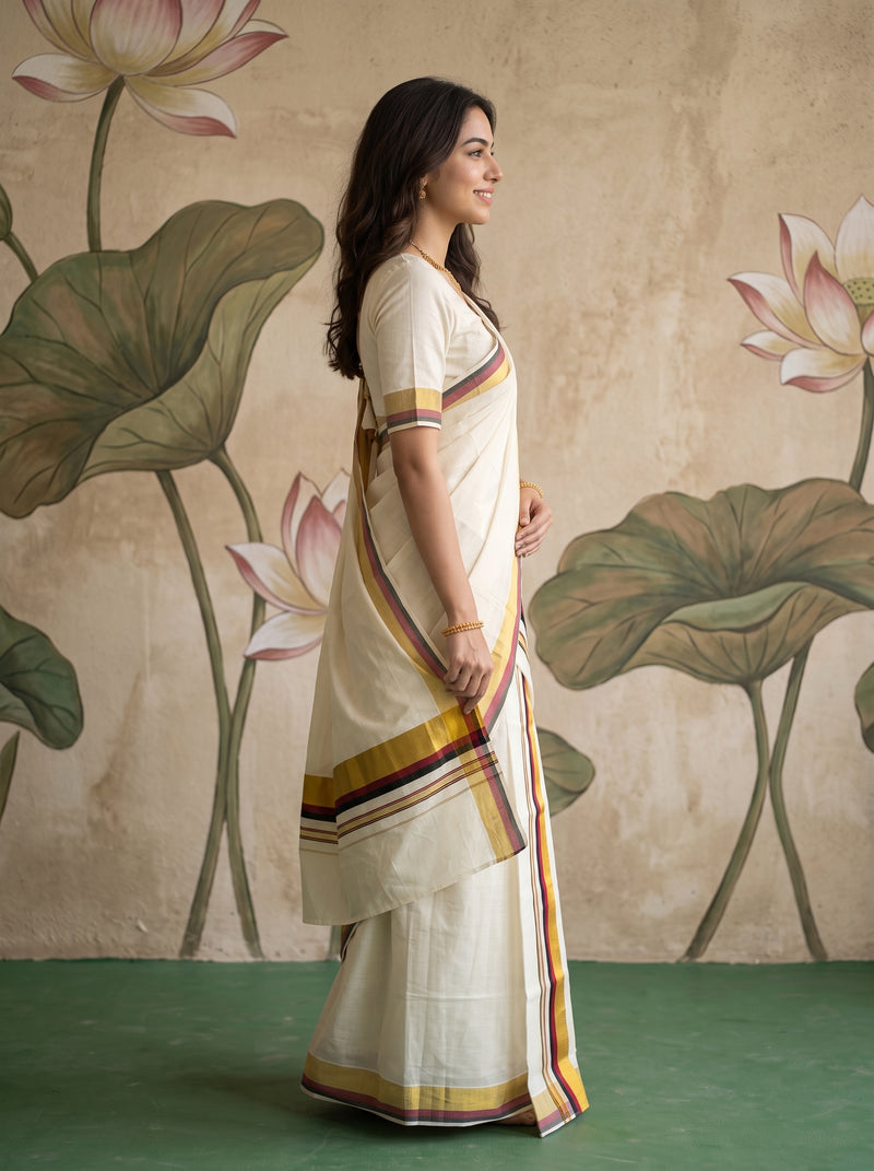 A woman wearing a cream-colored cotton saree with yellow, orange, and purple border stripes, standing in profile against a textured beige wall with painted lotus motifs.