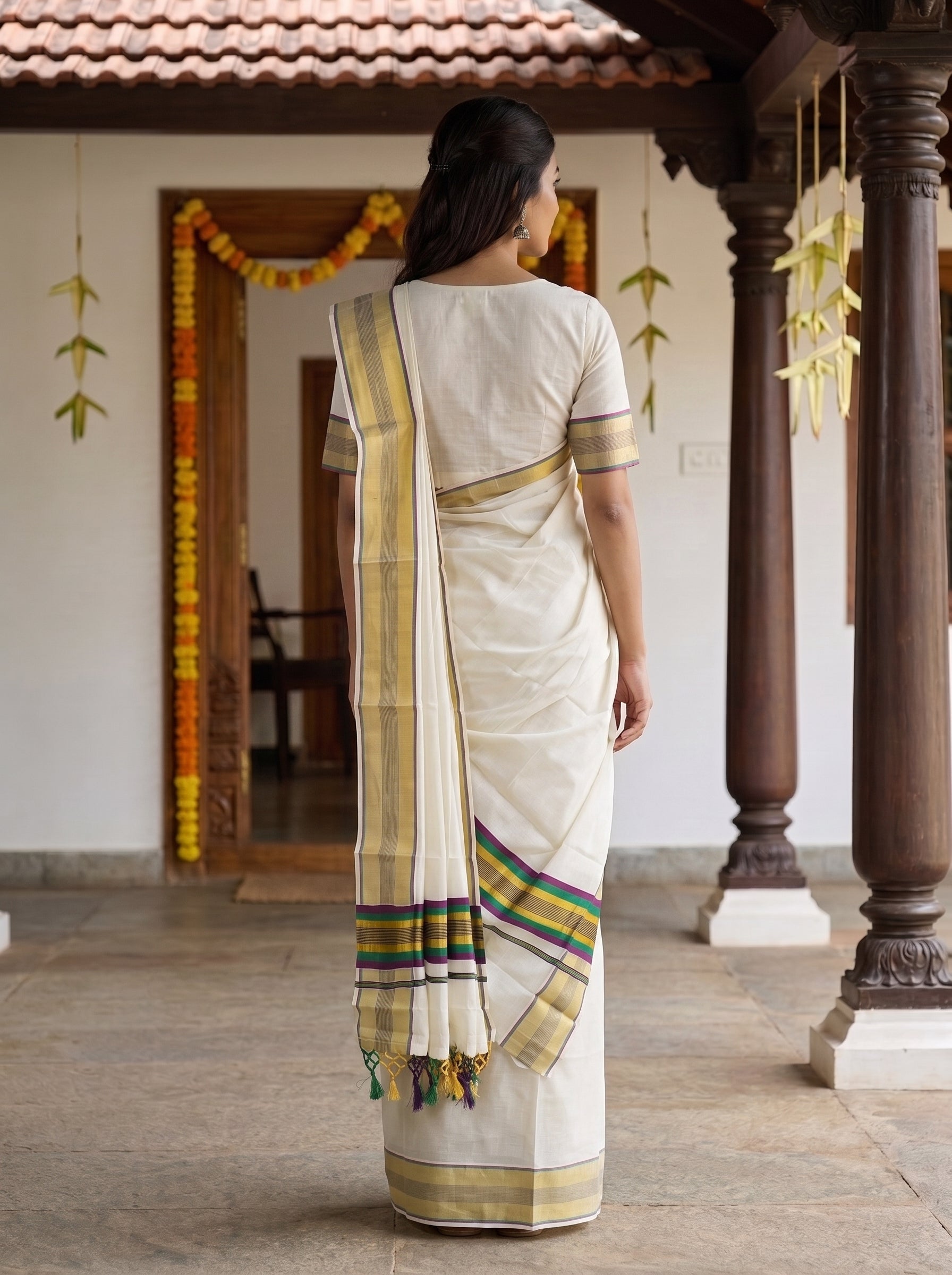 A woman viewed from behind wearing a cream-colored cotton saree with yellow, green, and blue striped borders, standing in a traditional courtyard with wooden pillars.