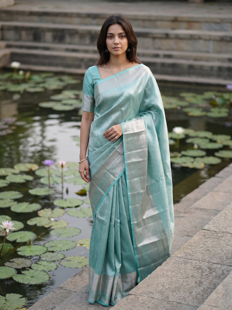 A woman wearing a light teal-green saree with a silver border and matching blouse stands on stone steps beside a pond with lily pads.