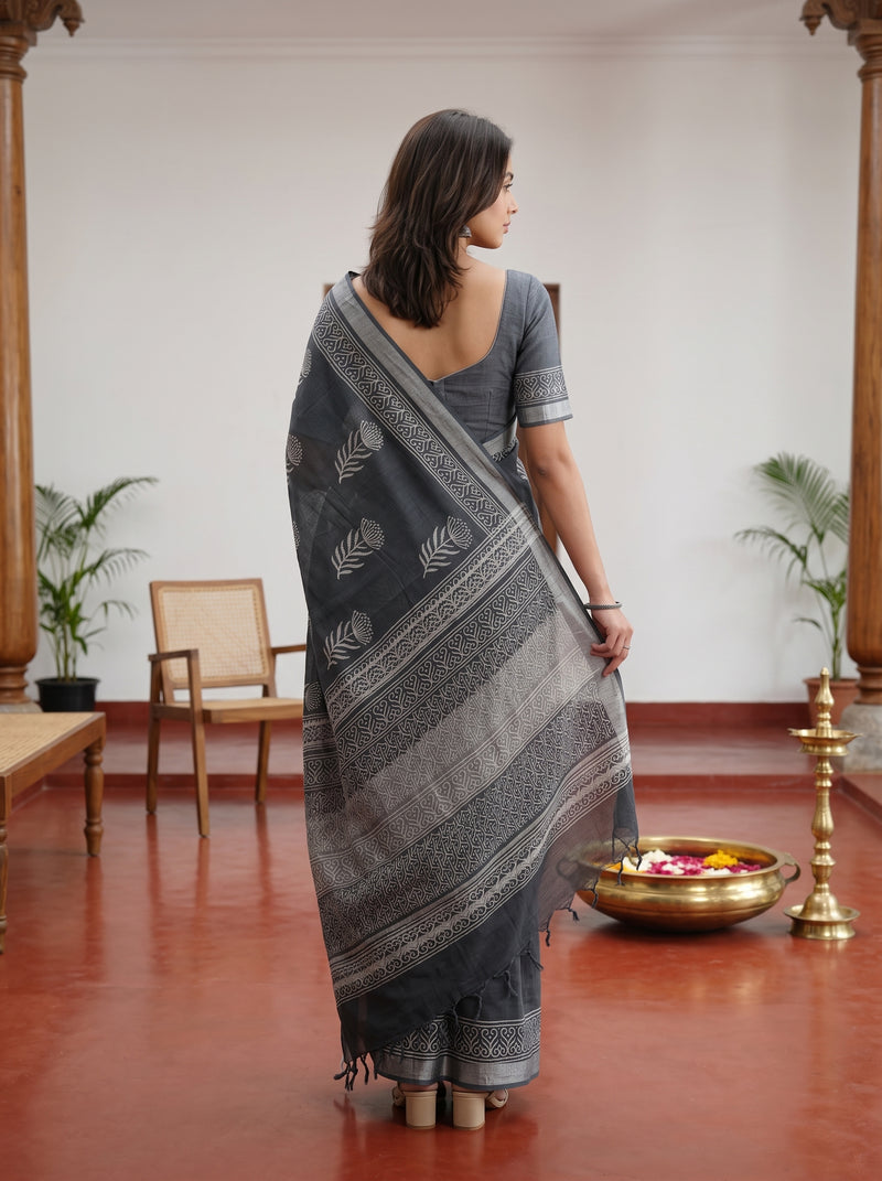 A woman stands with her back to the camera wearing a dark gray and silver patterned saree with a matching short-sleeve blouse in a traditional indoor setting with a red floor and wooden pillars.