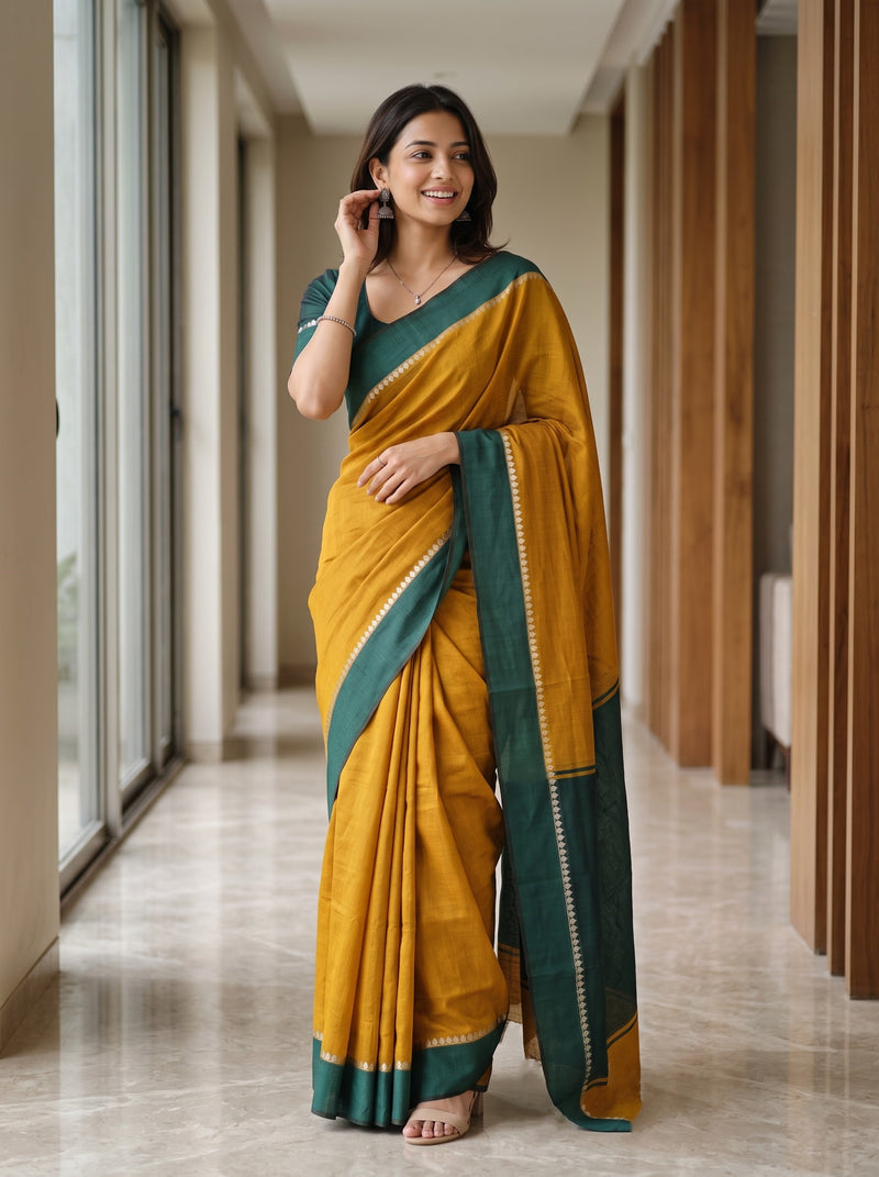 A woman wearing a mustard yellow and dark green saree with gold zari detailing, standing in a modern hallway with large windows and wooden slat walls.