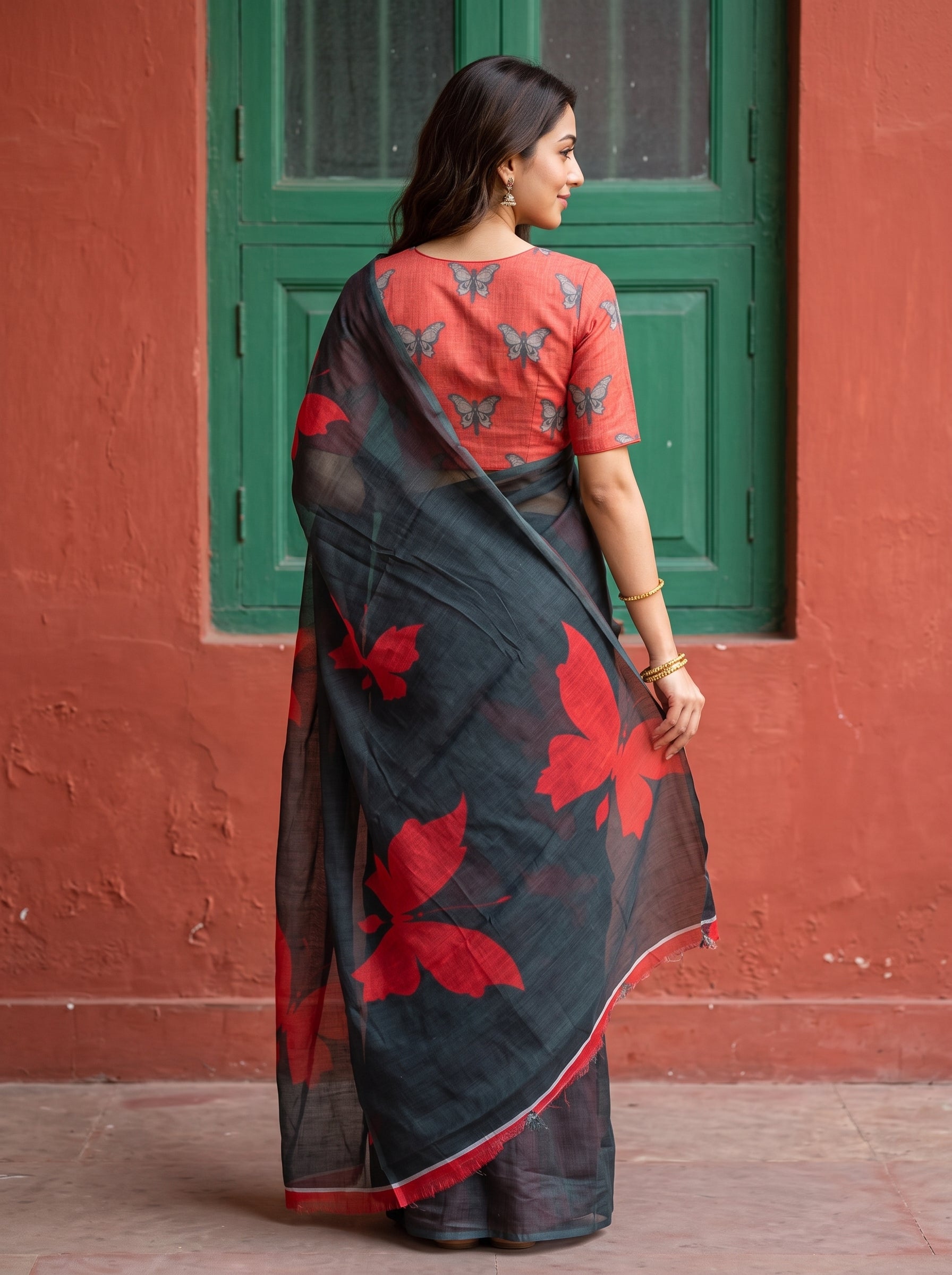 A woman wearing a black and red floral printed saree with a red printed blouse, standing in front of a red wall and green window.