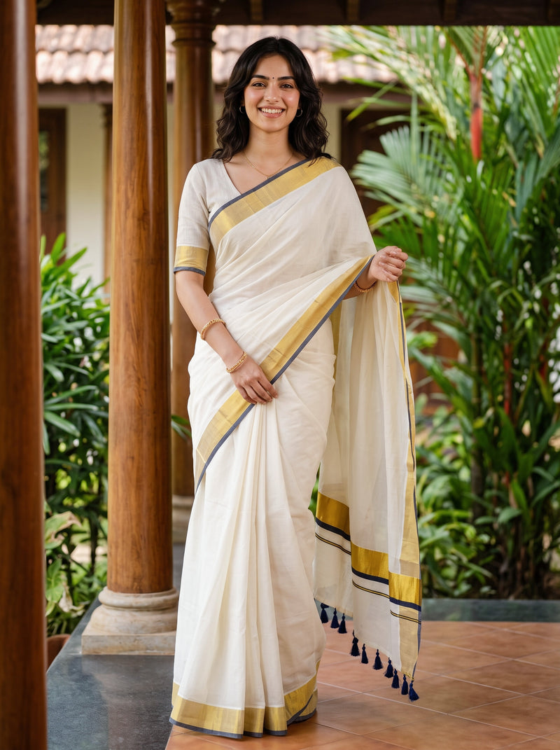 A woman wearing a cream-colored cotton saree with a gold and navy blue border, standing in a garden courtyard.