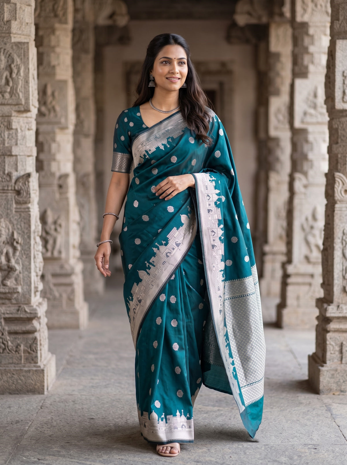 A woman wearing a teal blue and silver saree stands in a stone corridor with carved pillars. The saree has silver polka dots and a wide silver border with a white and silver patterned drape.