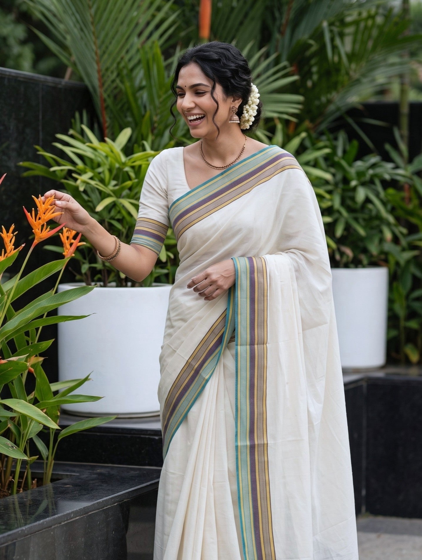 A woman wearing a cream-colored cotton saree with green, gold, and brown stripes on the border, paired with a matching cream blouse, posing in a garden setting next to orange flowers.