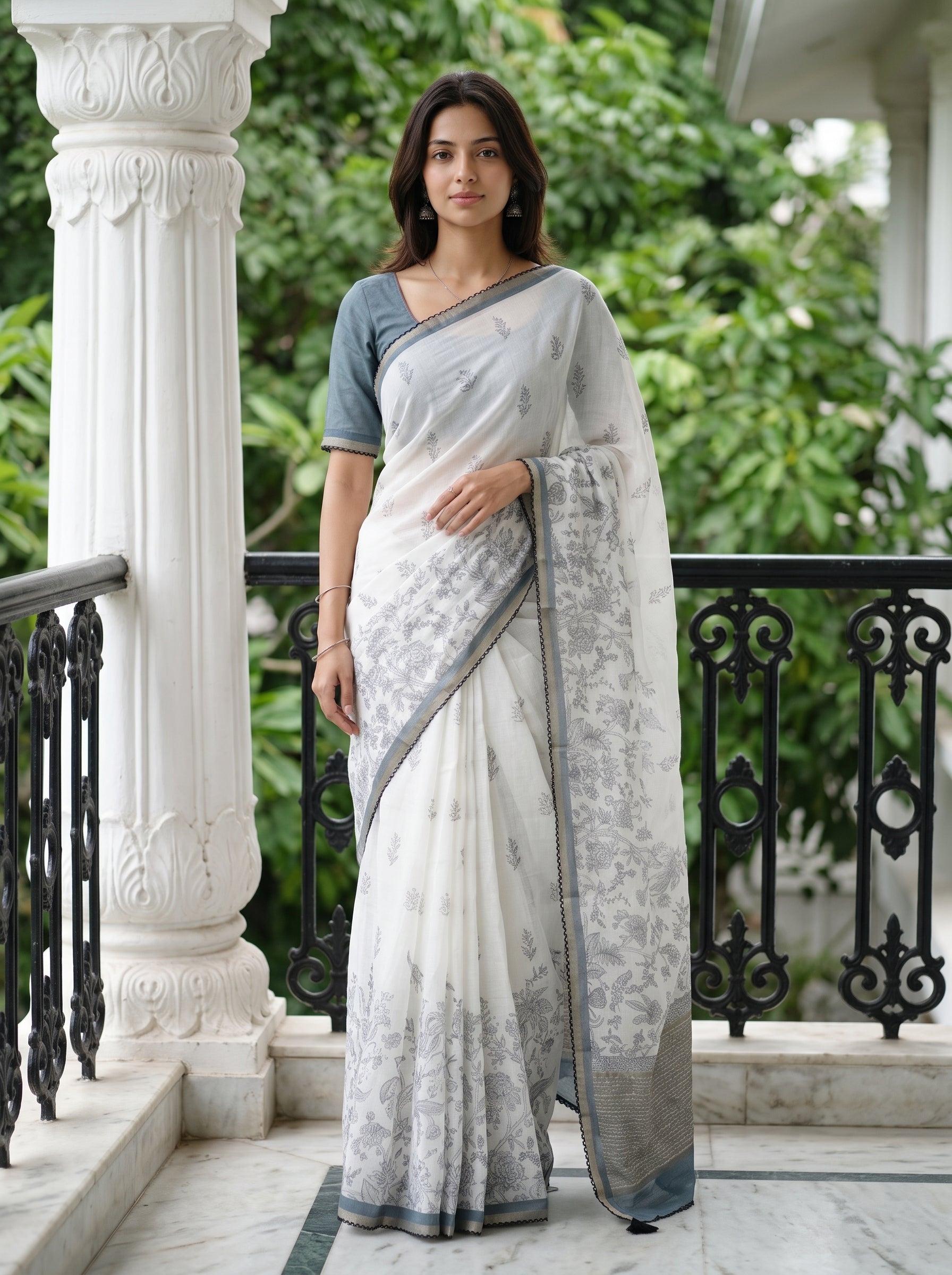 A woman wearing a white and gray patterned saree with a gray short-sleeve blouse, standing on a marble balcony with a white pillar and black railing behind her.