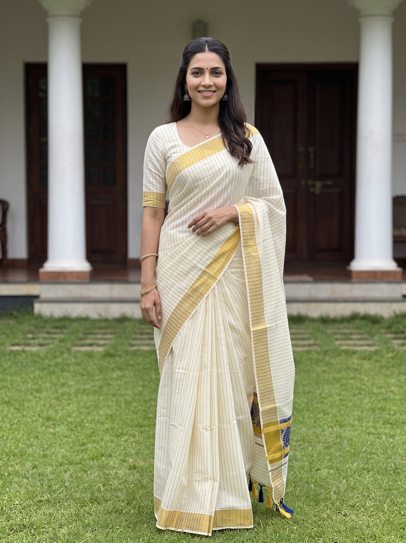A woman wearing a cream-colored saree with gold stripes and a matching cream blouse stands outdoors on a green lawn in front of a building with white columns and dark wooden doors.