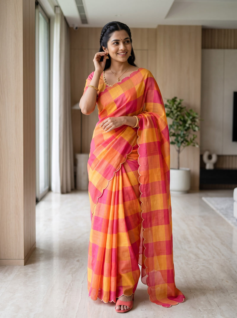 A woman wearing a bright orange and pink checkered saree stands in a modern indoor living space with light wood walls and a potted plant in the background.