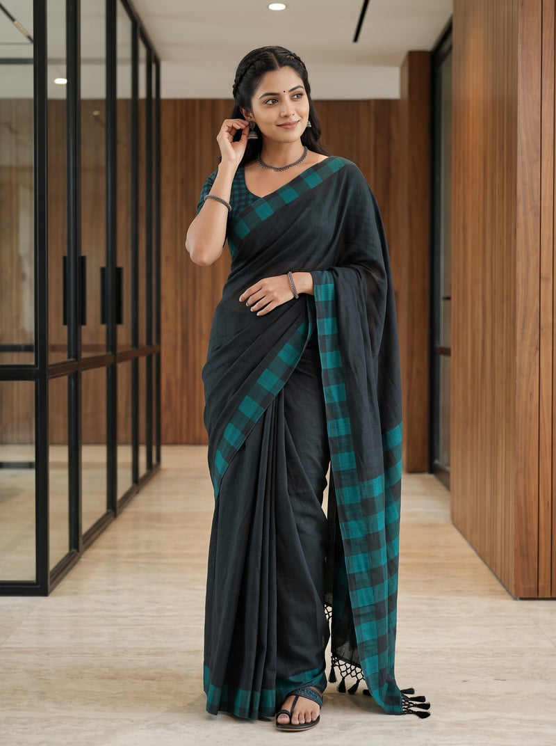 A woman modeling a black saree with a teal green and black checkered border, standing in a modern hallway with wood-paneled walls.