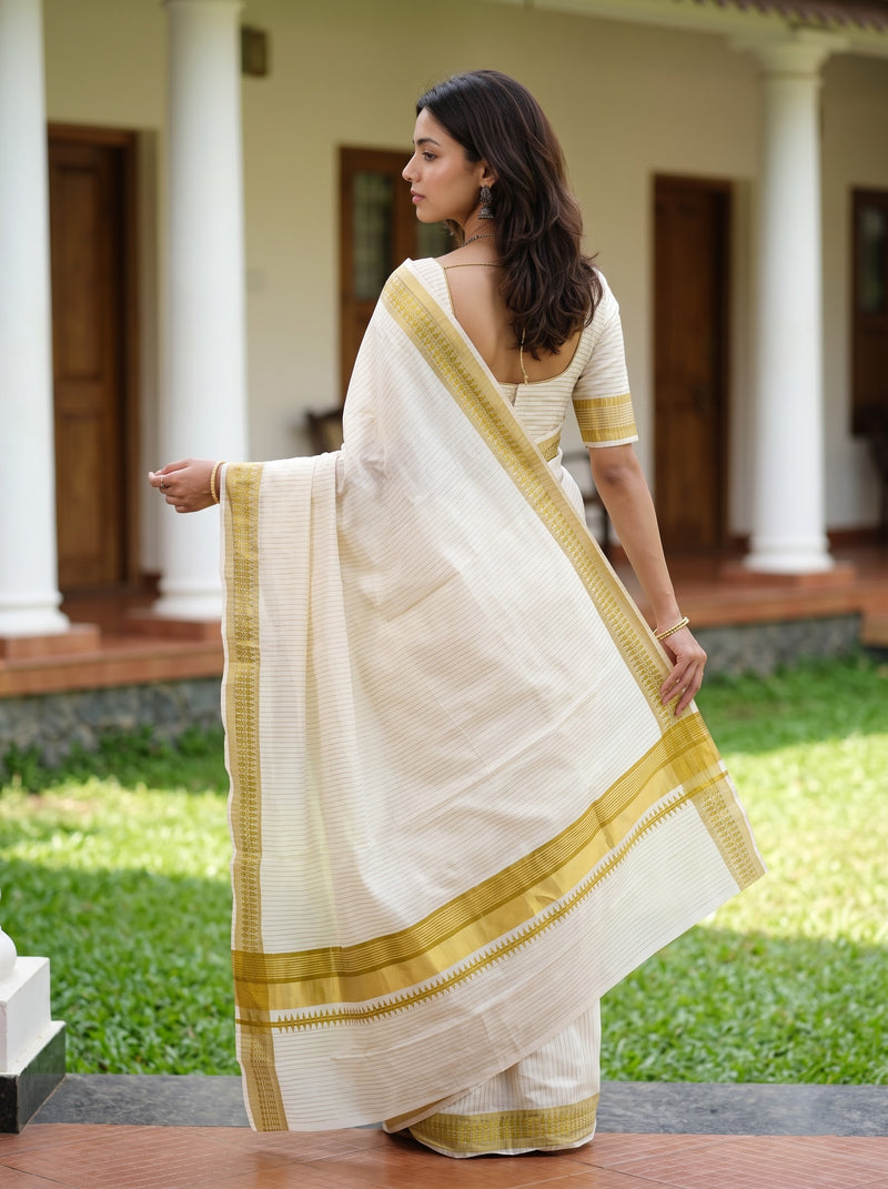 A woman wearing a cream-colored cotton saree with a gold border and matching cream blouse, standing outdoors in front of a building with white pillars and green grass.