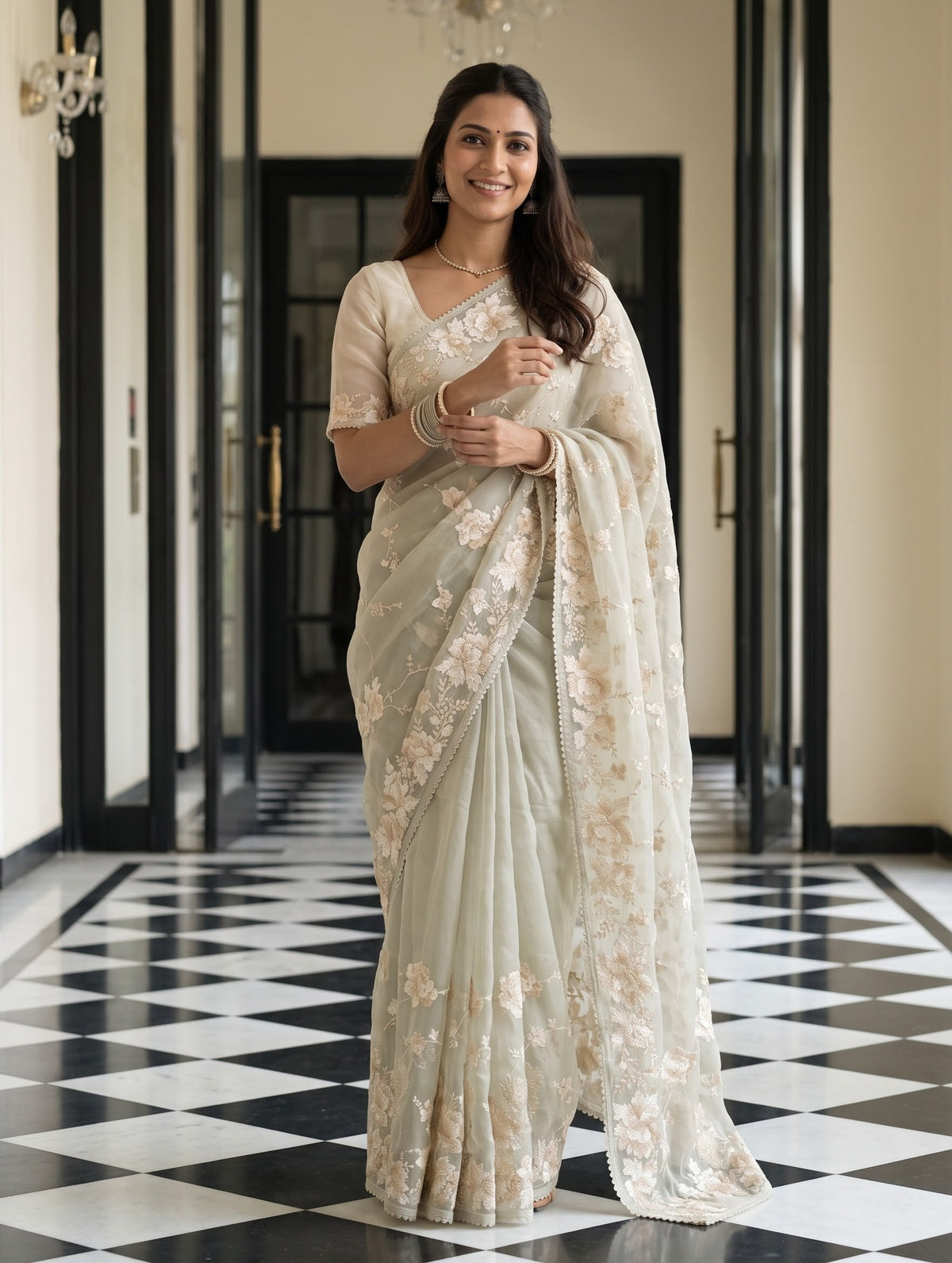 A woman stands in a hallway wearing a light beige floral saree with a matching blouse. The hallway features a black and white checkered floor and elegant cream-colored walls.