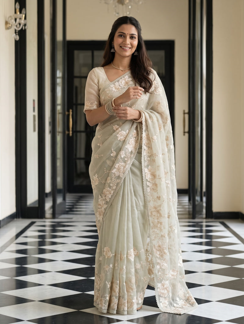 A woman stands in a hallway wearing a light beige floral saree with a matching blouse. The hallway features a black and white checkered floor and elegant cream-colored walls.