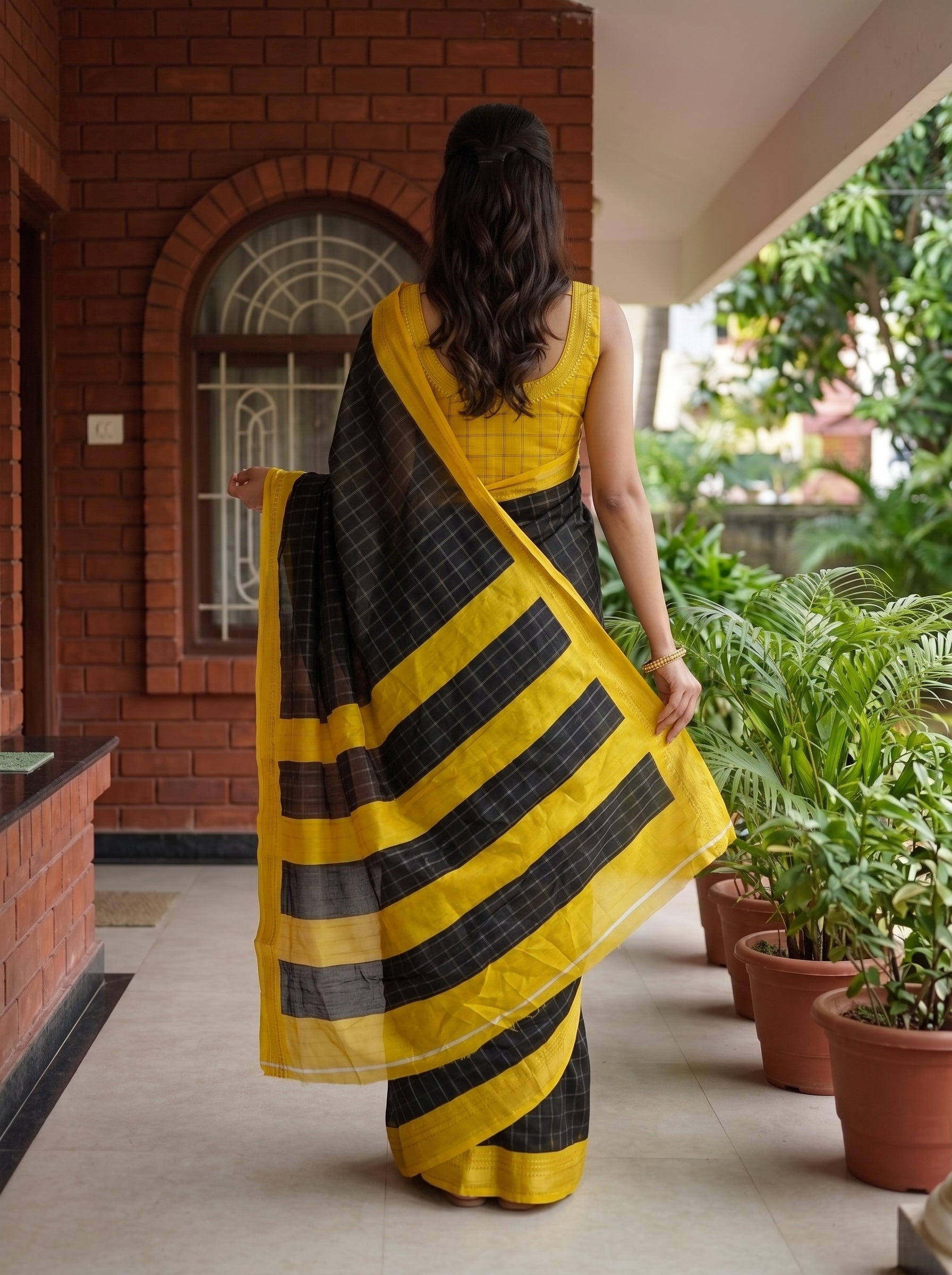 A woman viewed from behind wearing a yellow and black striped saree with a yellow sleeveless blouse, standing on a covered walkway beside potted plants.