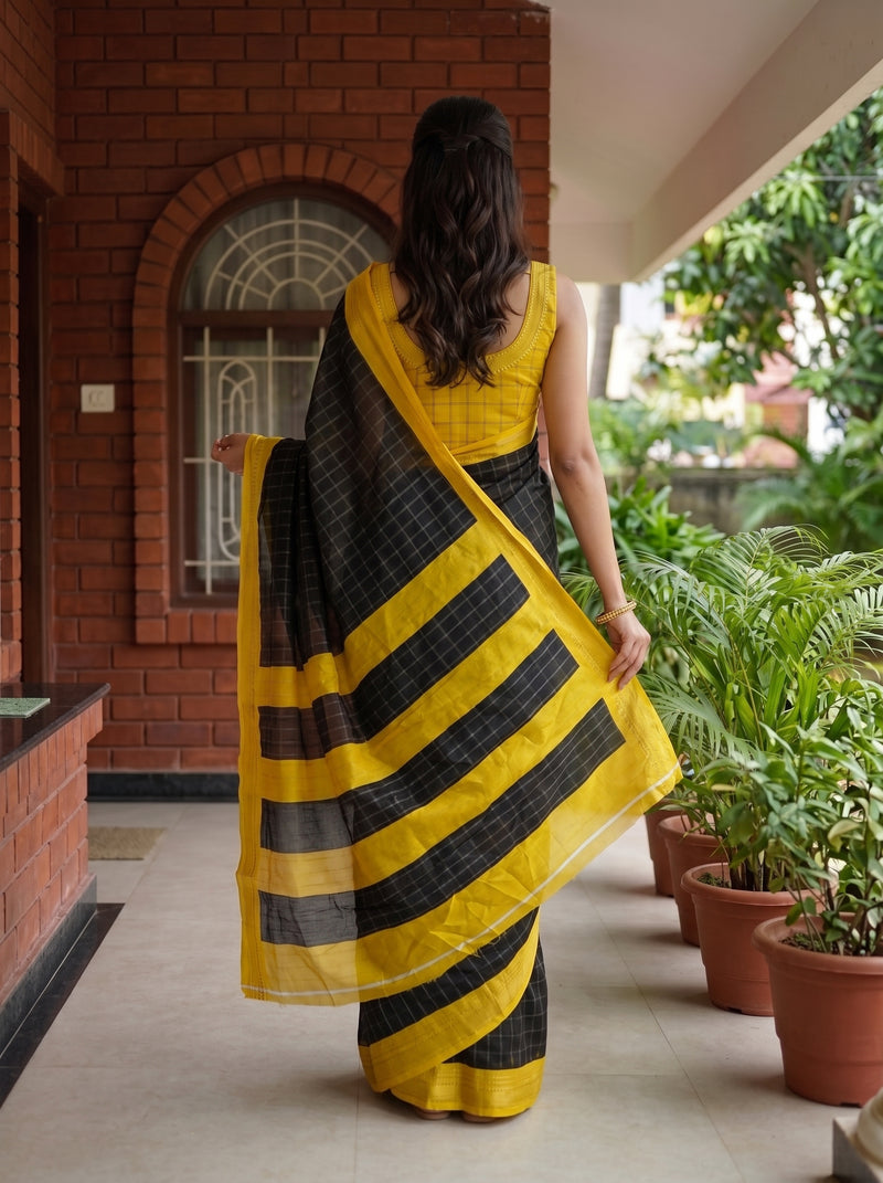 A woman viewed from behind wearing a yellow and black striped saree with a yellow sleeveless blouse, standing on a covered walkway beside potted plants.