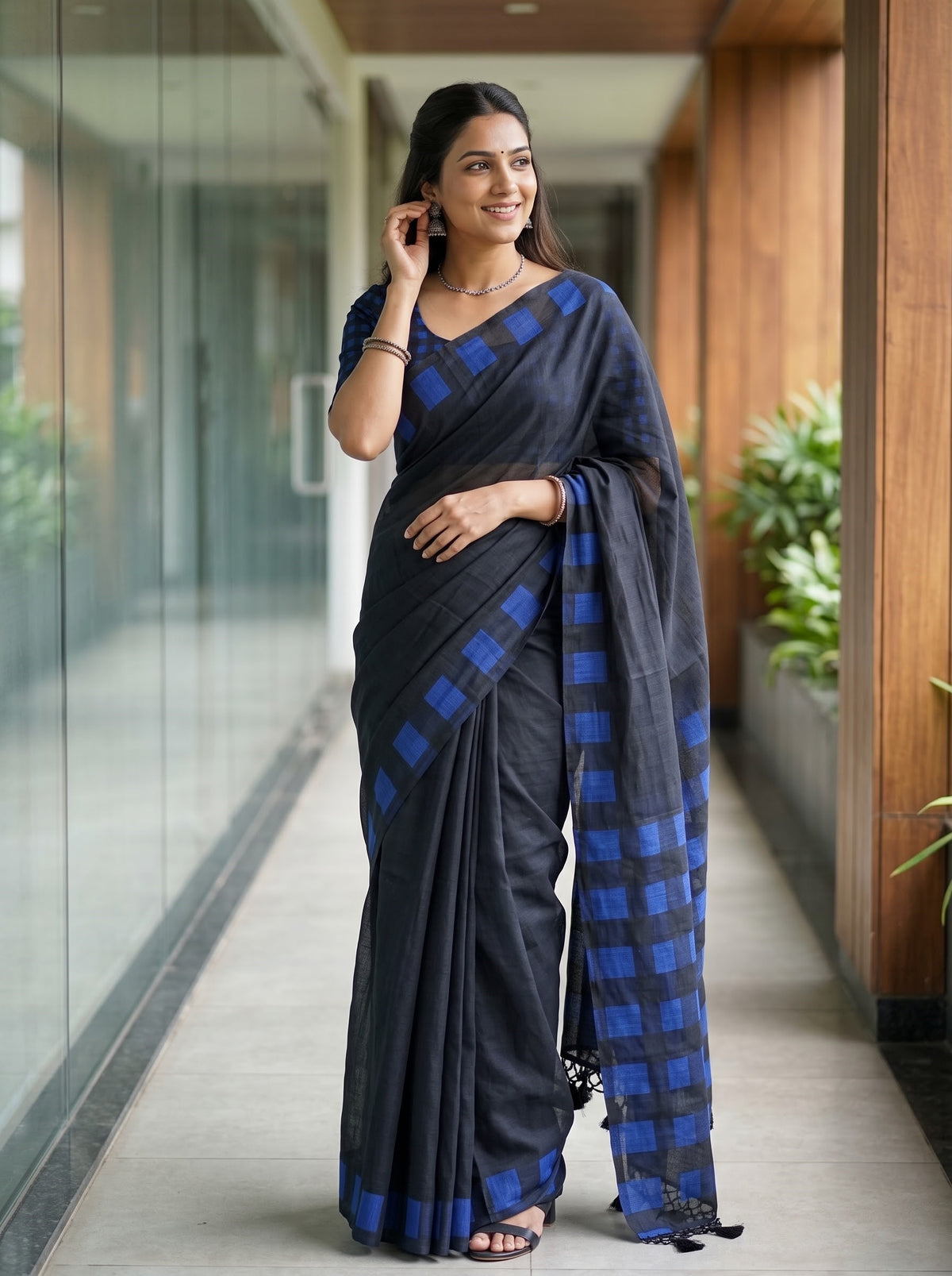 A woman modeling a dark gray and blue checkered saree in a modern indoor hallway with glass walls and wooden columns.