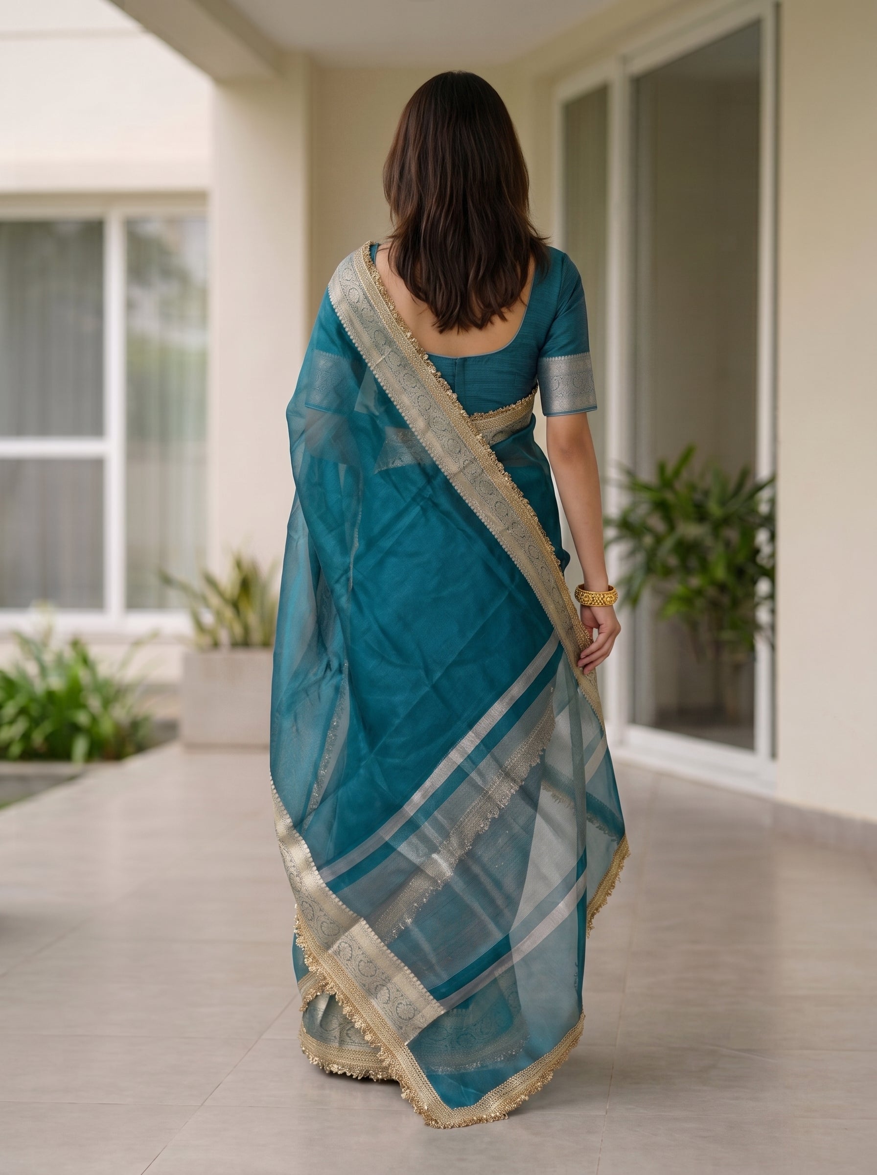 A woman viewed from behind wearing a teal blue saree with a silver and gold zari border, walking in a modern indoor corridor with plants and large windows.