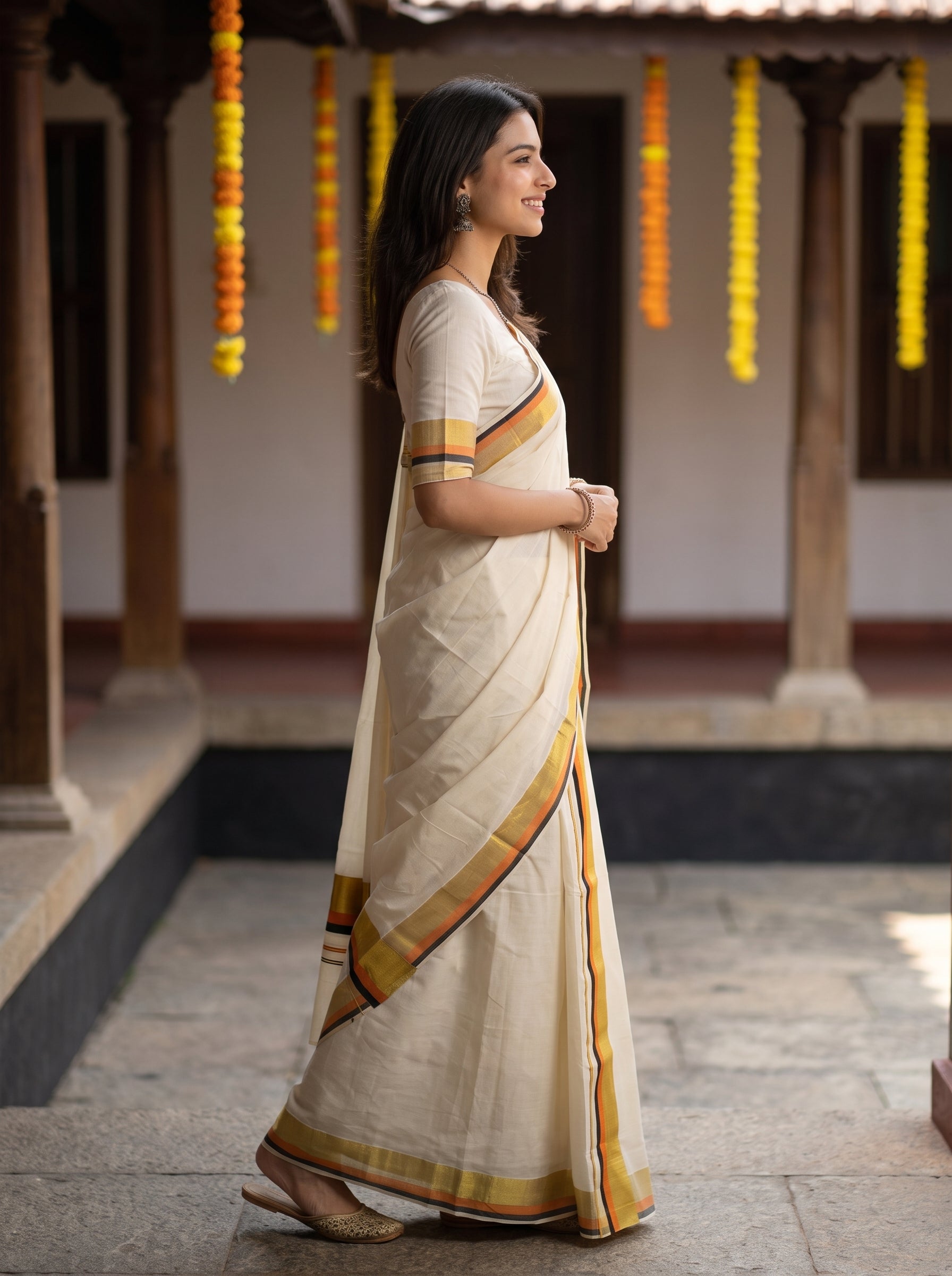 A woman wearing a cream-colored cotton saree with yellow, orange, and red border stripes, standing in profile in a traditional courtyard.