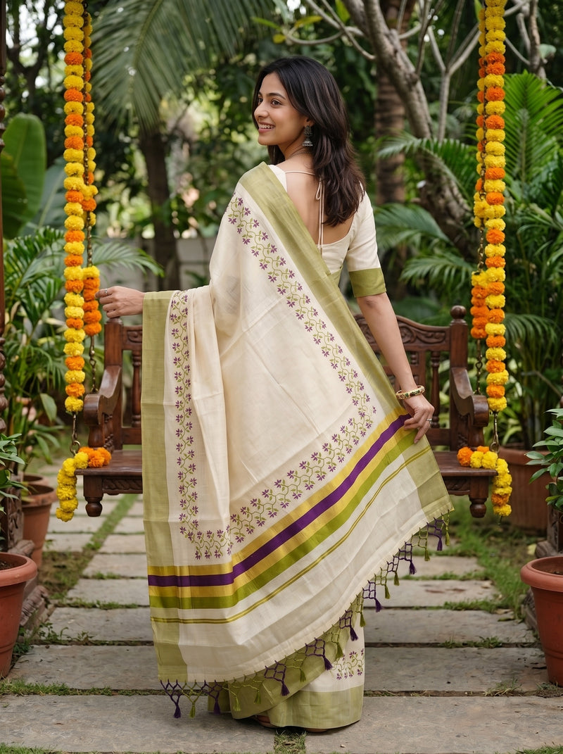 A woman wearing a light beige and olive green saree with purple and yellow stripes, standing in a garden courtyard decorated with hanging marigold garlands.