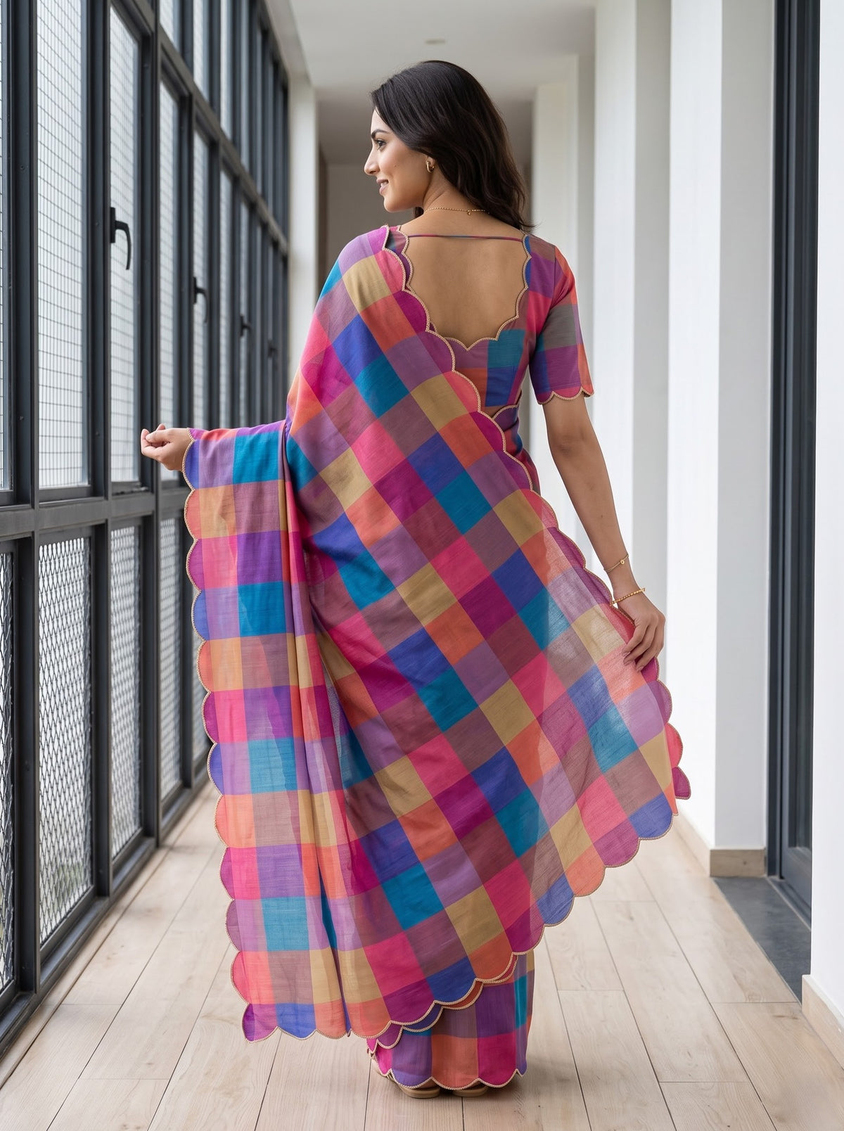 A woman wearing a vibrant multicolor checkered saree with a scalloped border and a matching short-sleeve blouse, standing in a modern indoor hallway.