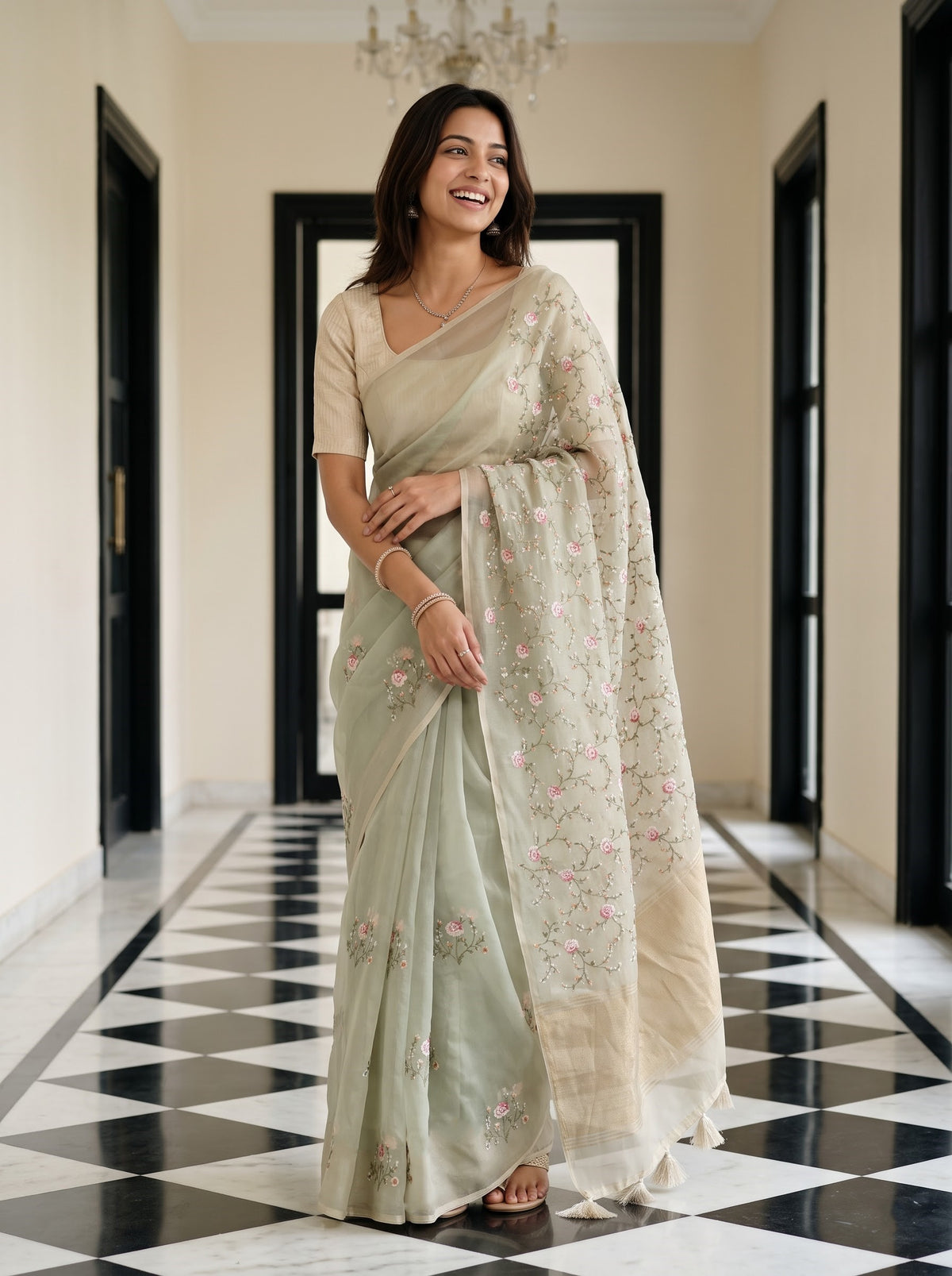 A woman wearing a light sage green saree with pink floral embroidery and a matching embroidered dupatta, standing in a hallway with a black and white checkered floor.