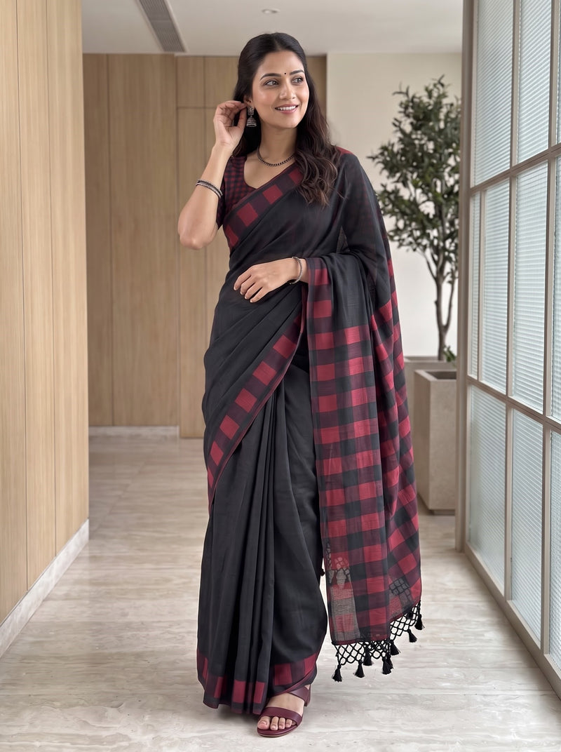 A woman modeling a black saree with a red and black checkered border and pallu, paired with a matching red and black checkered blouse, standing in a modern indoor hallway.