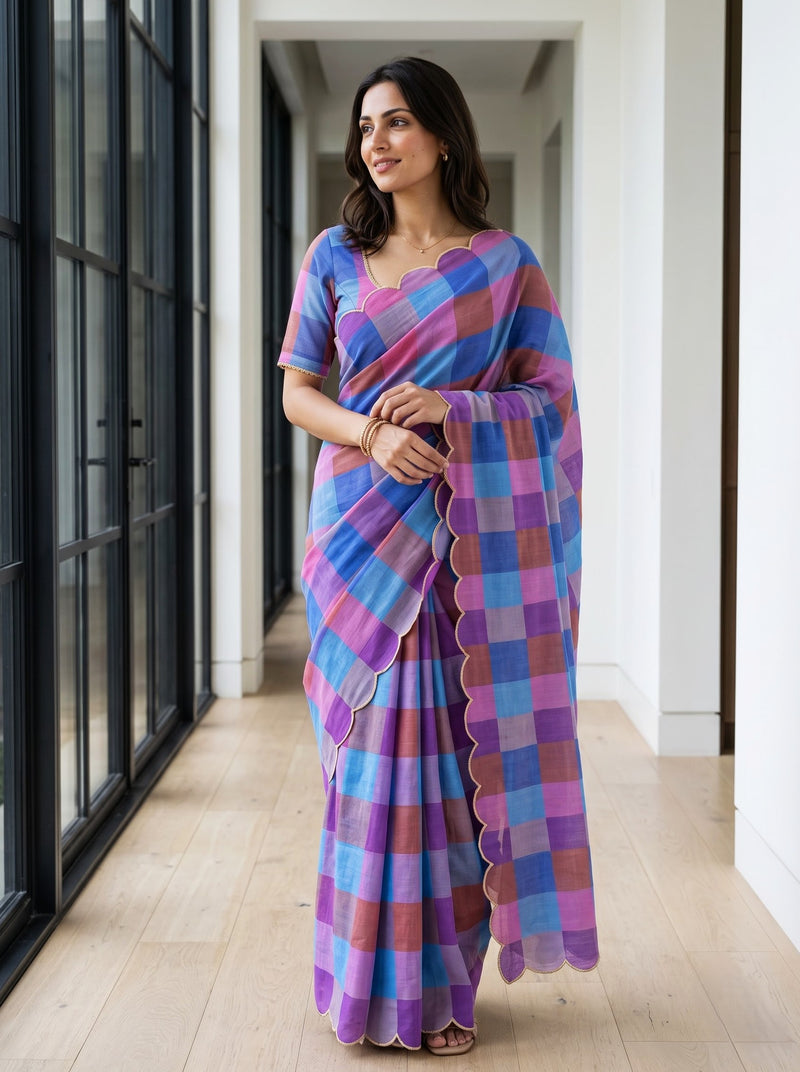 A woman modeling a vibrant checkered saree with a scalloped border, featuring shades of blue, purple, and pink, standing in a modern hallway with light wood floors.