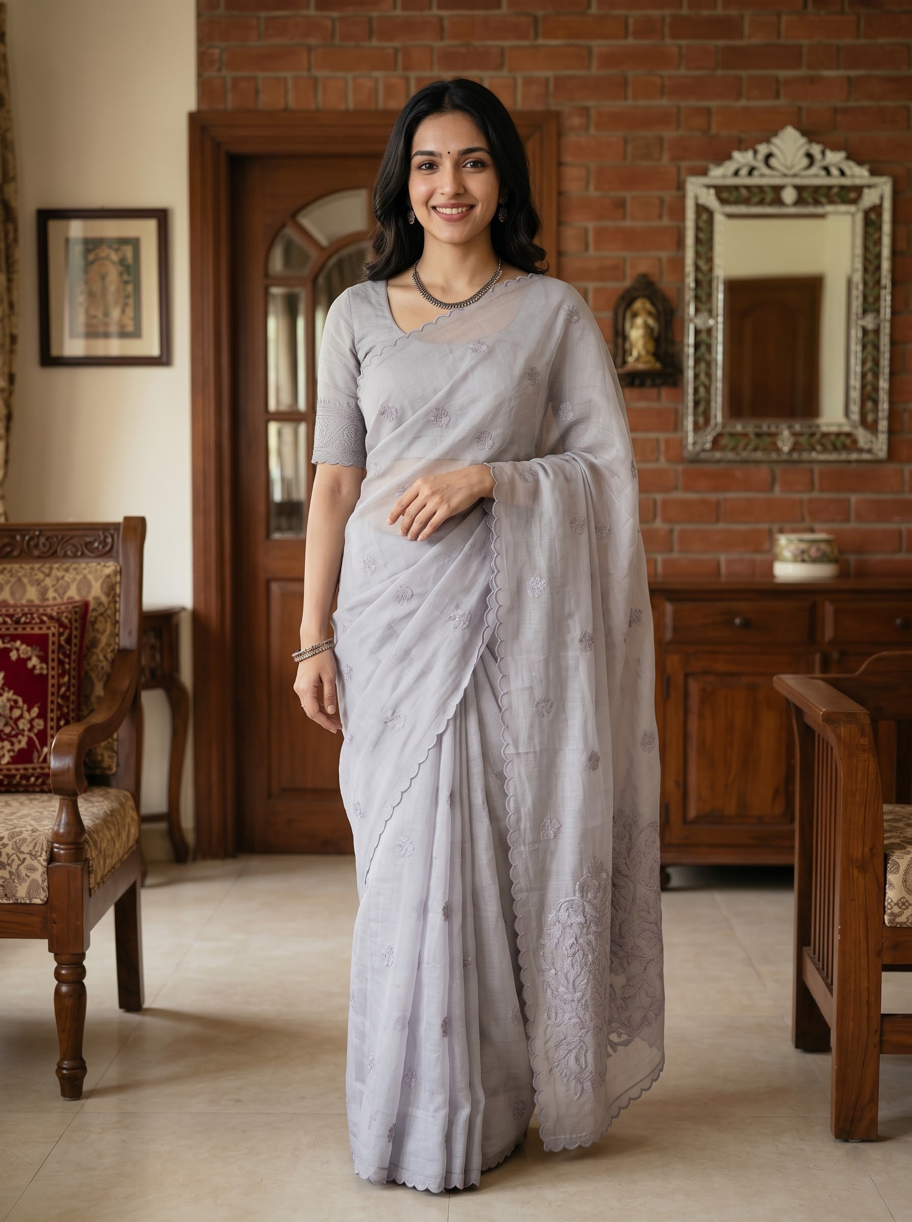 A woman wearing a light gray saree with a matching short-sleeve blouse, standing in a traditional indoor room with a brick wall, wooden furniture, and a mirror.