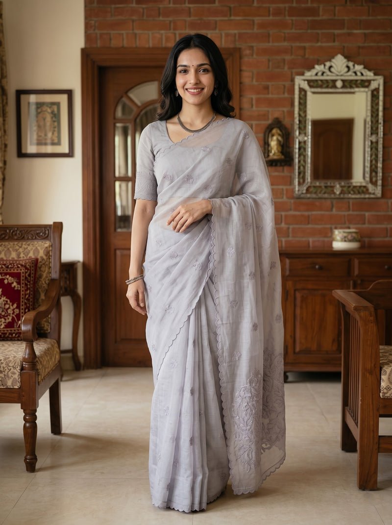 A woman wearing a light gray saree with a matching short-sleeve blouse, standing in a traditional indoor room with a brick wall, wooden furniture, and a mirror.
