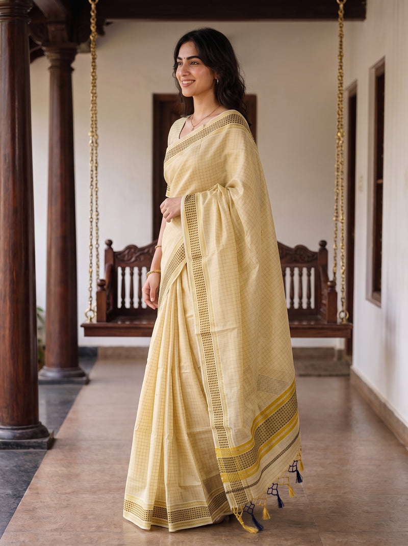A woman wearing a light yellow checkered saree with a yellow and black border, standing in a traditional indoor corridor with wooden pillars and hanging swings.
