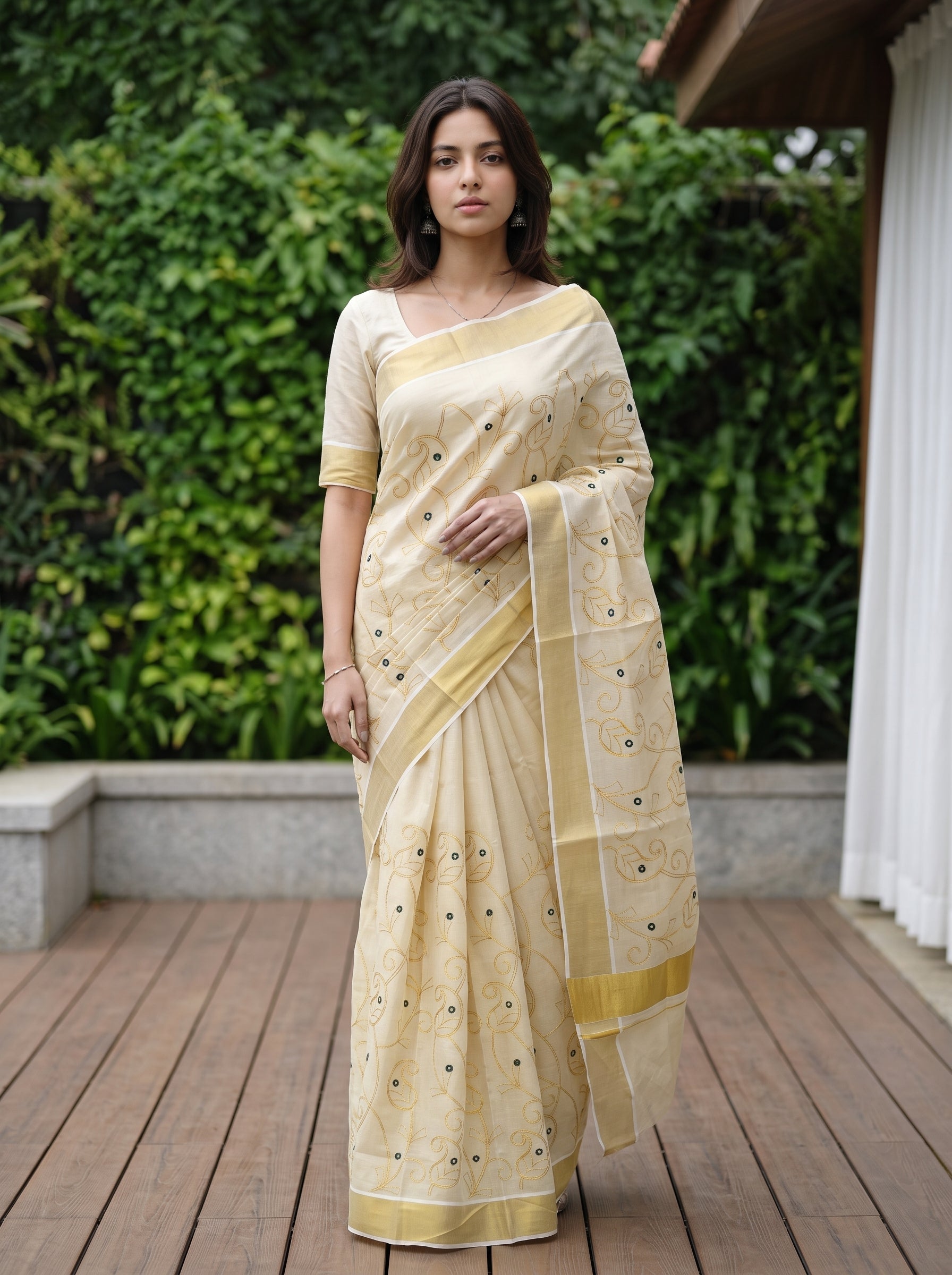 A woman wearing a light beige and yellow saree with gold embroidery and a matching blouse, standing on a wooden deck with a green garden background.