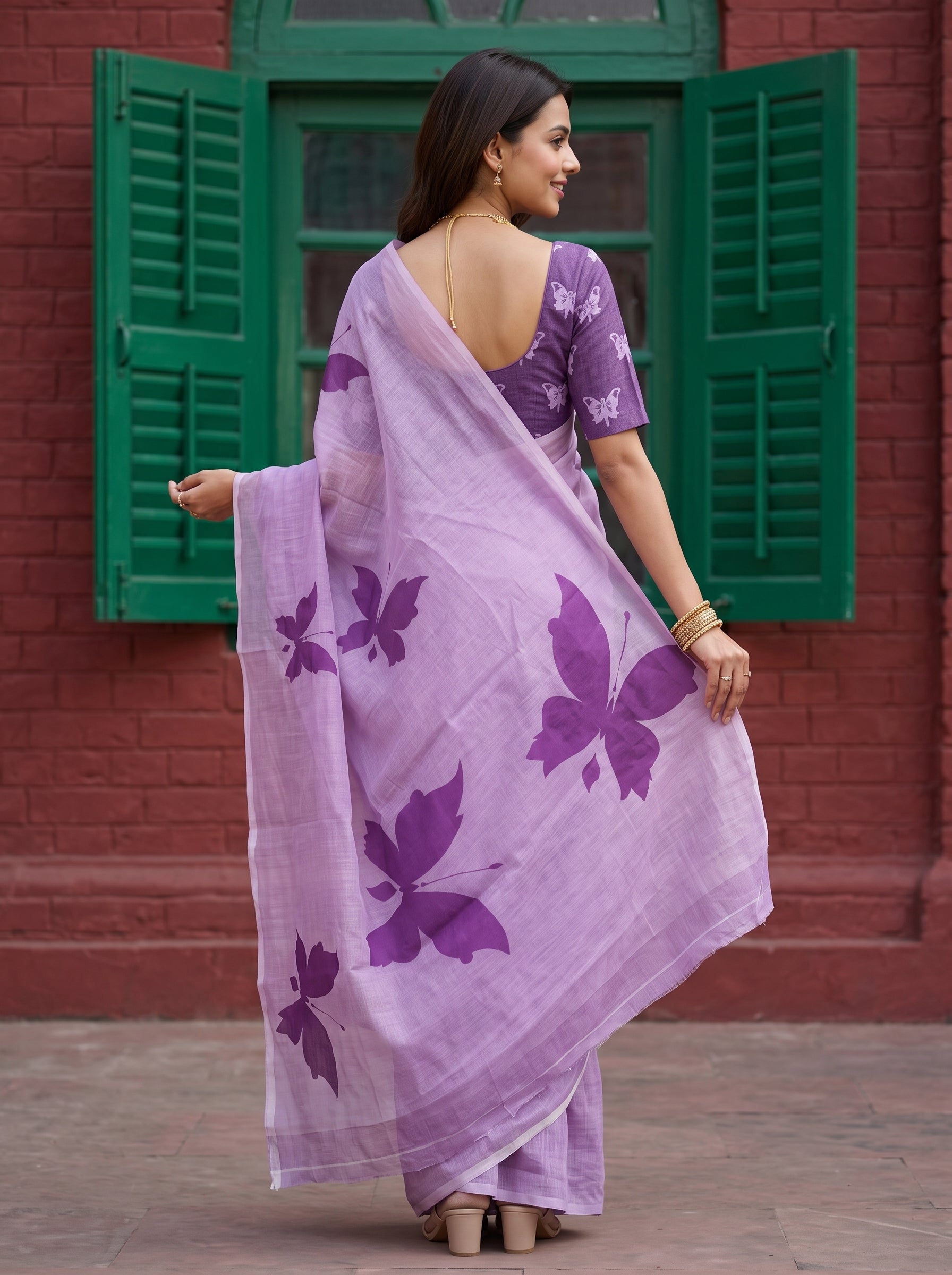 A woman wearing a lavender purple saree with large dark purple butterfly prints, paired with a matching purple floral blouse, standing in front of a red brick wall with green shutters.