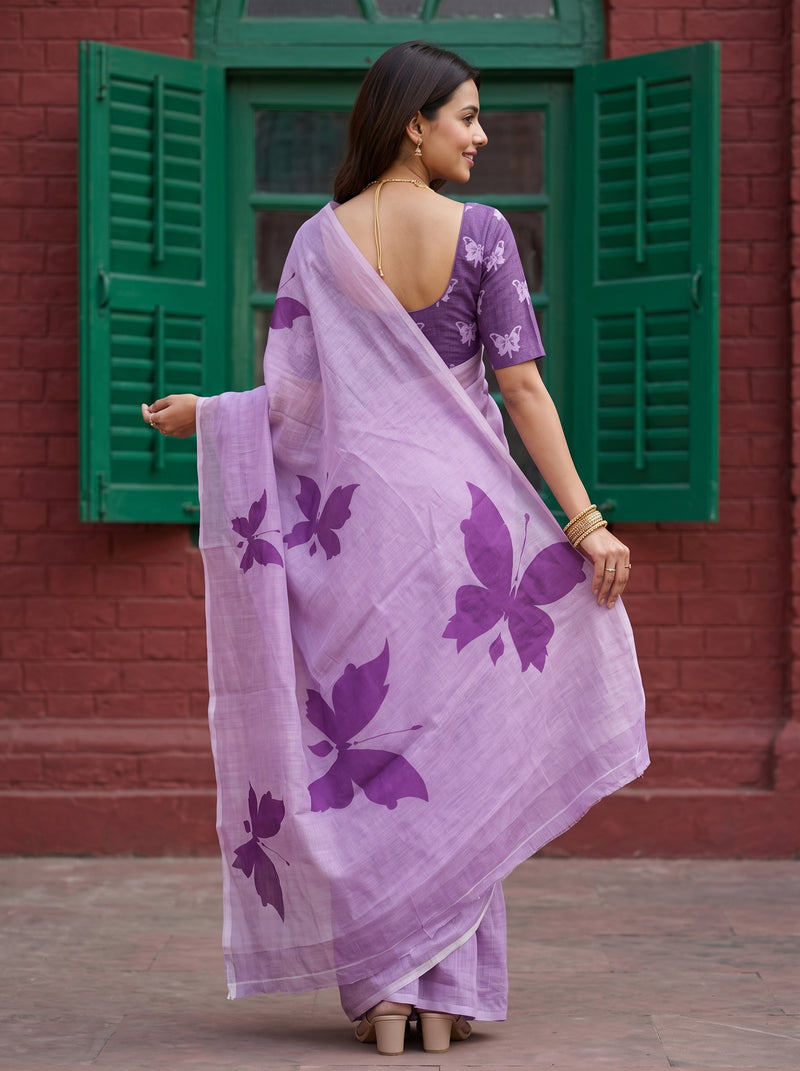 A woman wearing a lavender purple saree with large dark purple butterfly prints, paired with a matching purple floral blouse, standing in front of a red brick wall with green shutters.