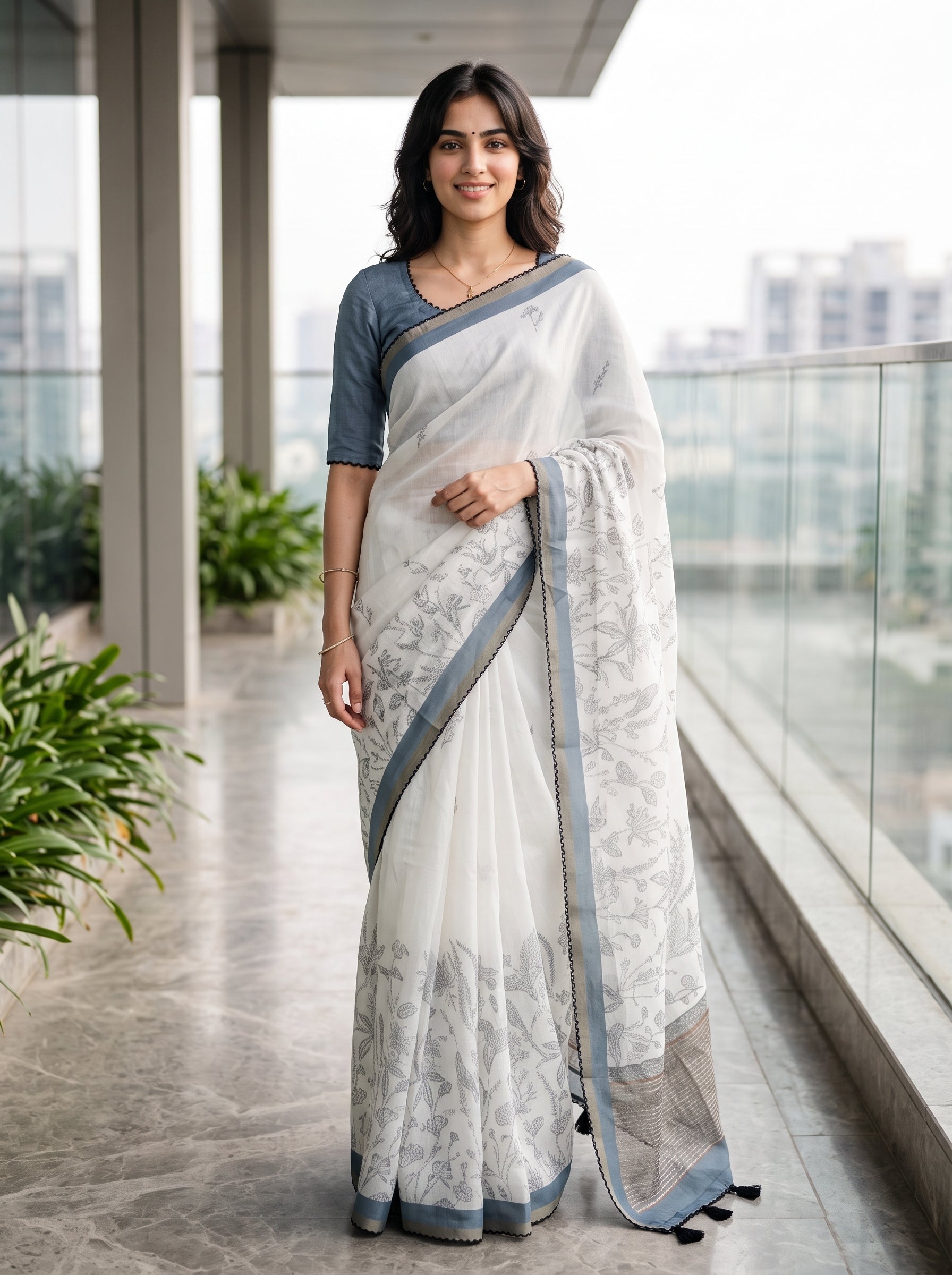 A woman stands on a modern balcony wearing a white saree with light gray floral patterns, a dusty blue sleeveless blouse, and a matching blue border.