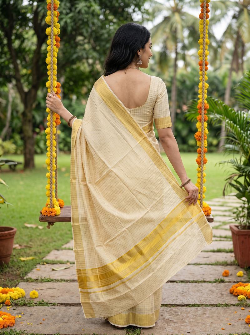 A woman wearing a cream-colored checkered saree with a yellow border and a matching cream blouse stands on a stone path in a garden, holding a swing decorated with yellow and orange marigold garlands.