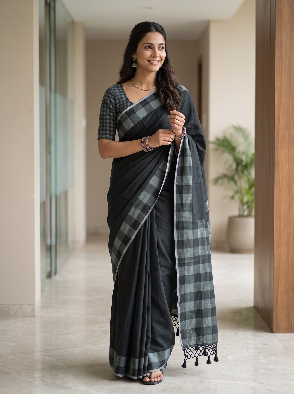 A woman modeling a black and gray plaid saree with a matching checkered blouse, standing in a modern indoor hallway with beige walls and tiled floors.