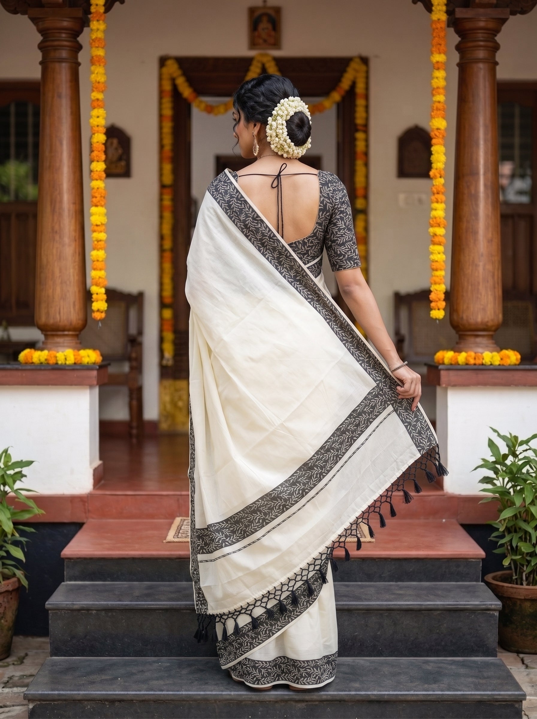 A woman stands on steps in front of a decorated doorway wearing a cream/off-white saree with a black patterned border and a matching black patterned blouse. The scene includes wooden pillars, yellow flower garlands, and potted plants.