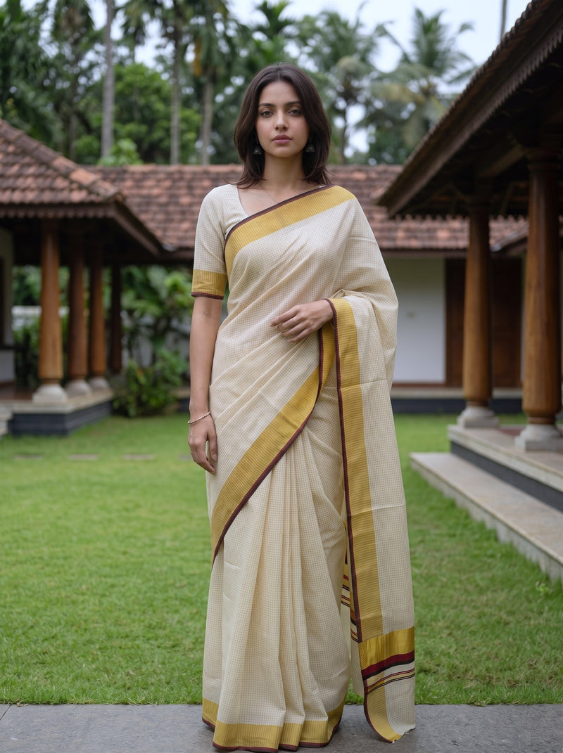 A woman stands outdoors in a traditional cream-colored saree with mustard yellow and maroon borders, set against a background of green grass, trees, and wooden pavilion structures.