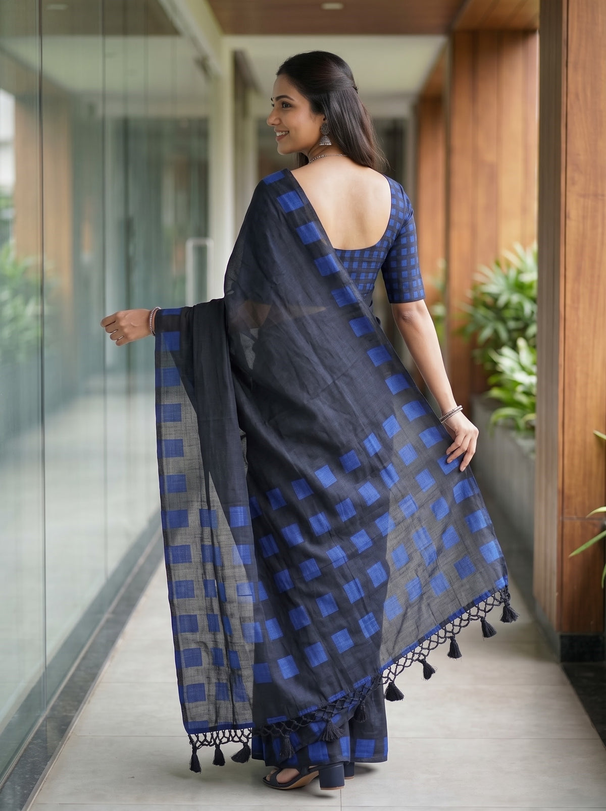 A woman wearing a dark gray and blue checkered saree with a matching blue checkered blouse, standing in a modern indoor hallway with glass walls and wooden columns.