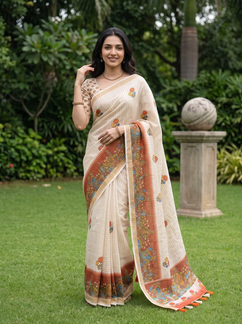 A woman wearing a cream-colored saree with orange, blue, and green floral patterns stands in a lush green garden. The saree features a matching printed blouse and a decorative border with tassels.