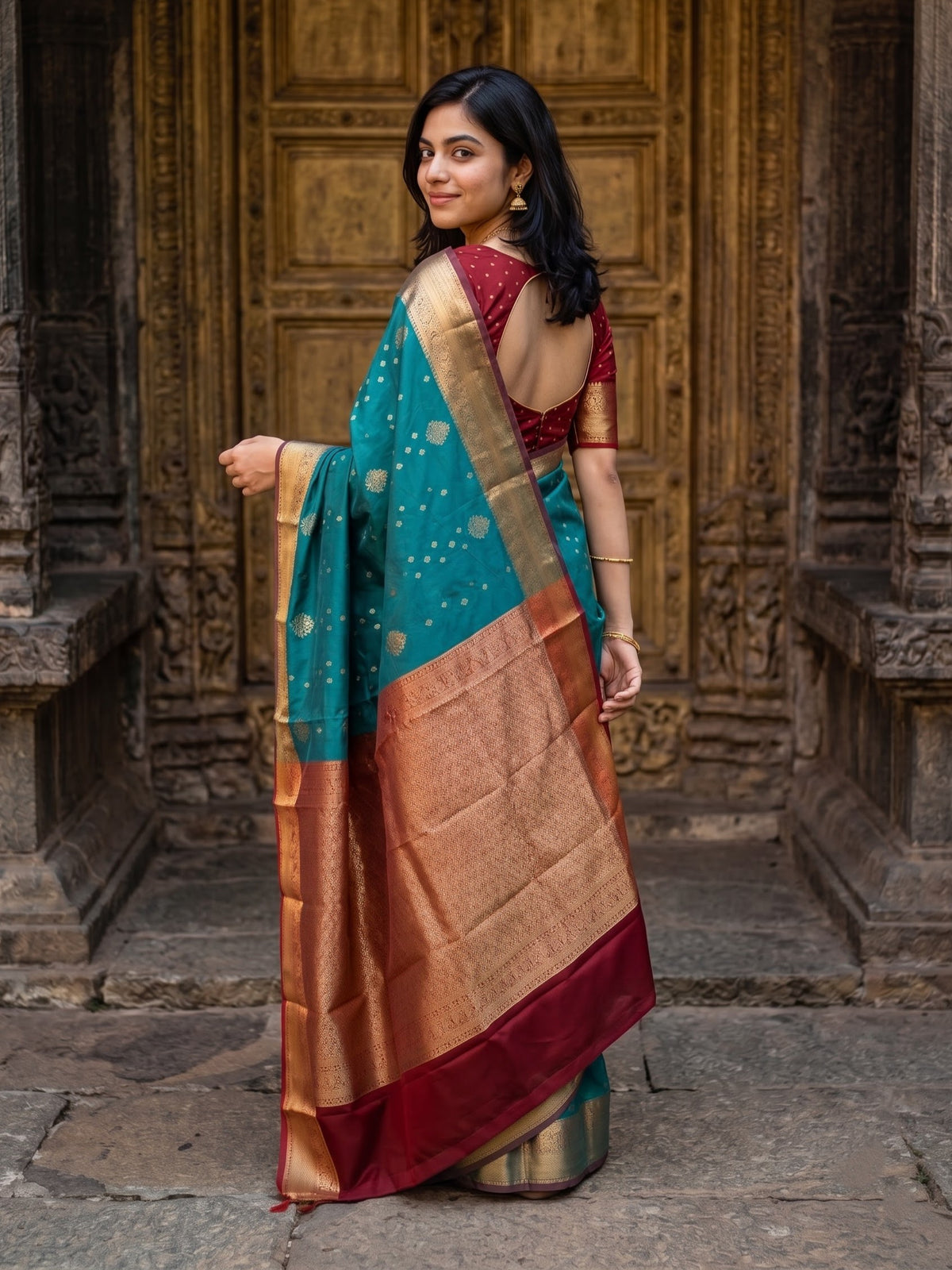 A woman wearing a teal and gold zari saree with a deep red border and a maroon embroidered blouse, standing in front of an ornate carved wooden door.