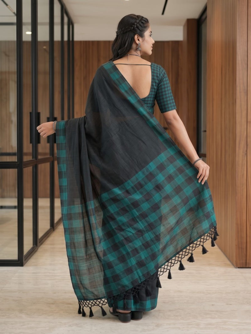 A woman wearing a black and teal green plaid saree with a matching checkered blouse, standing in a modern indoor hallway with wood-paneled walls and glass partitions.