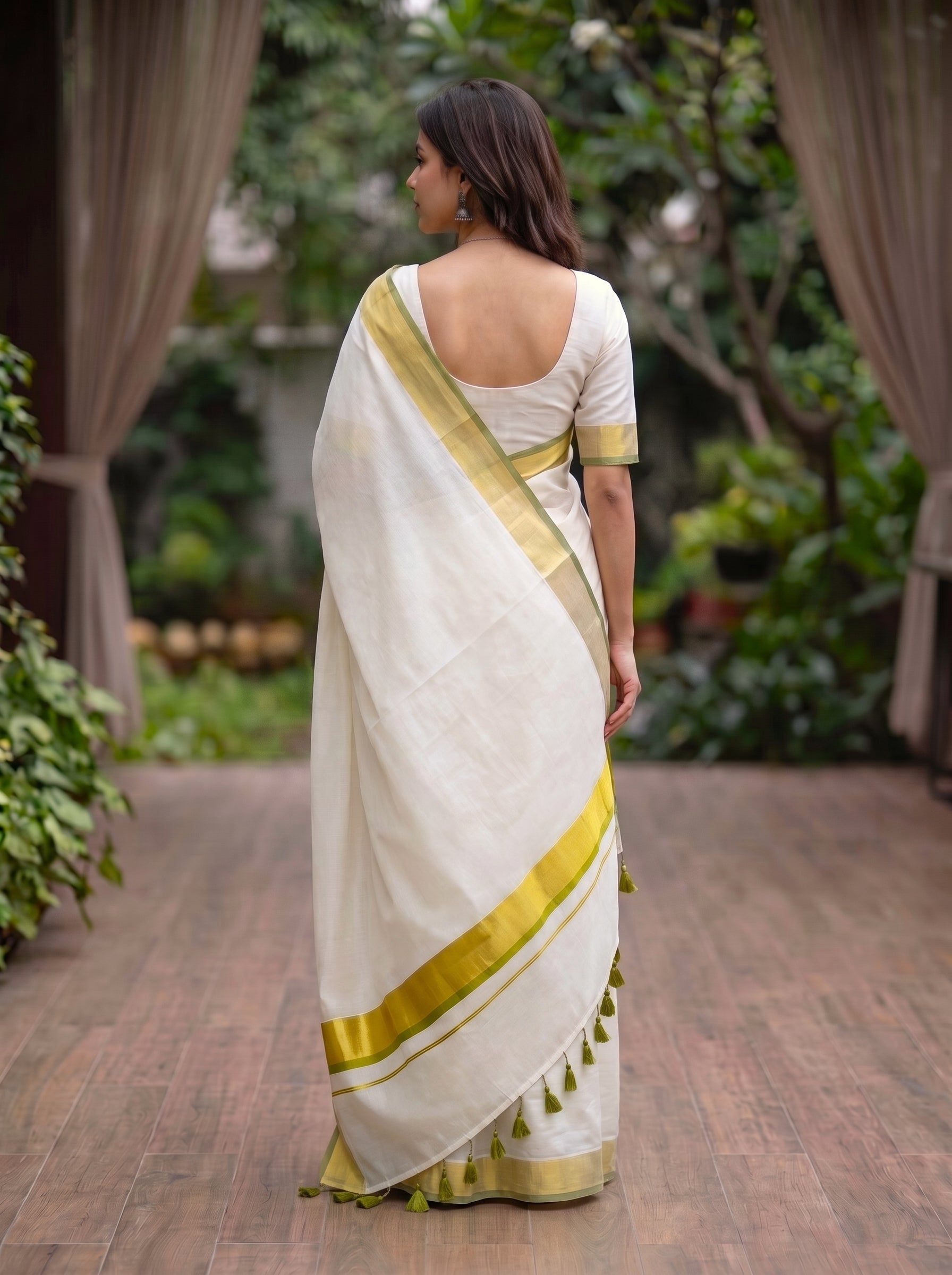 A woman wearing a white silk saree with a gold border and green tassels, shown from the back in an outdoor garden setting.