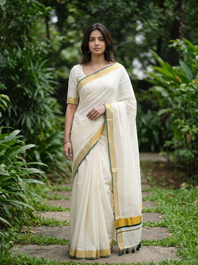 A woman wearing a cream-colored saree with gold and green border details, standing on a stone path surrounded by lush green garden foliage.