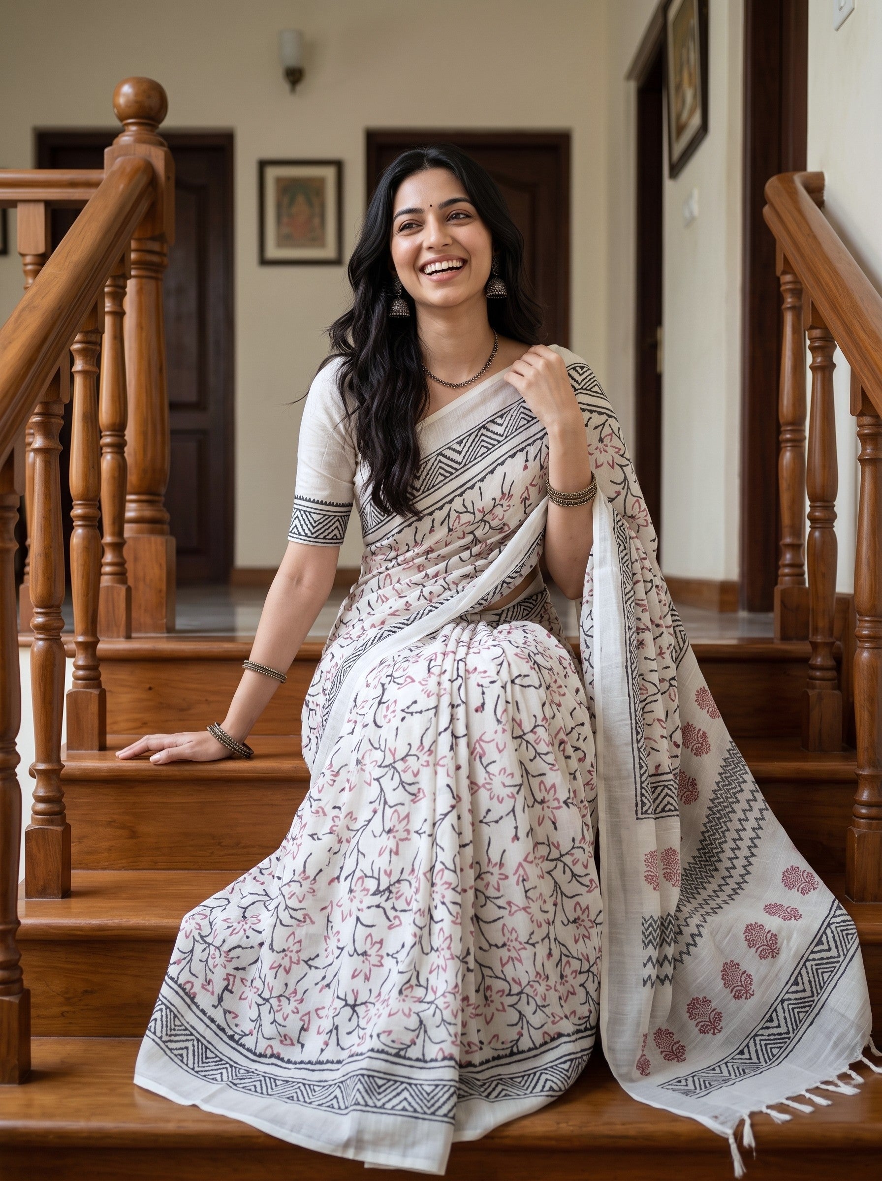 A woman wearing a white printed saree with black and pink floral patterns sits on wooden stairs in a home interior.